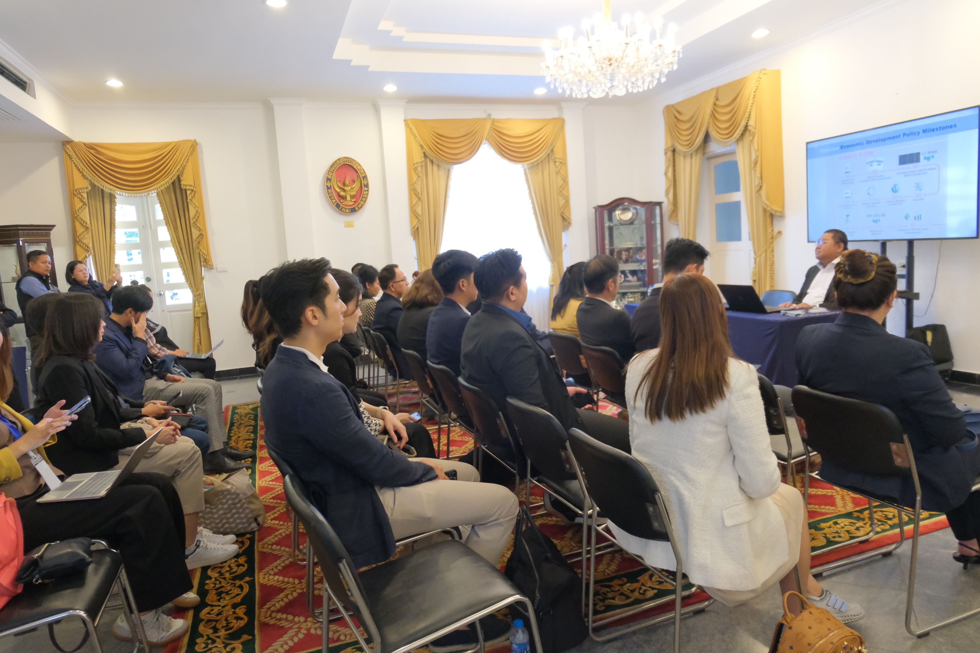 Audience listens attentively to a speaker presenting data on a large screen in a formal room.