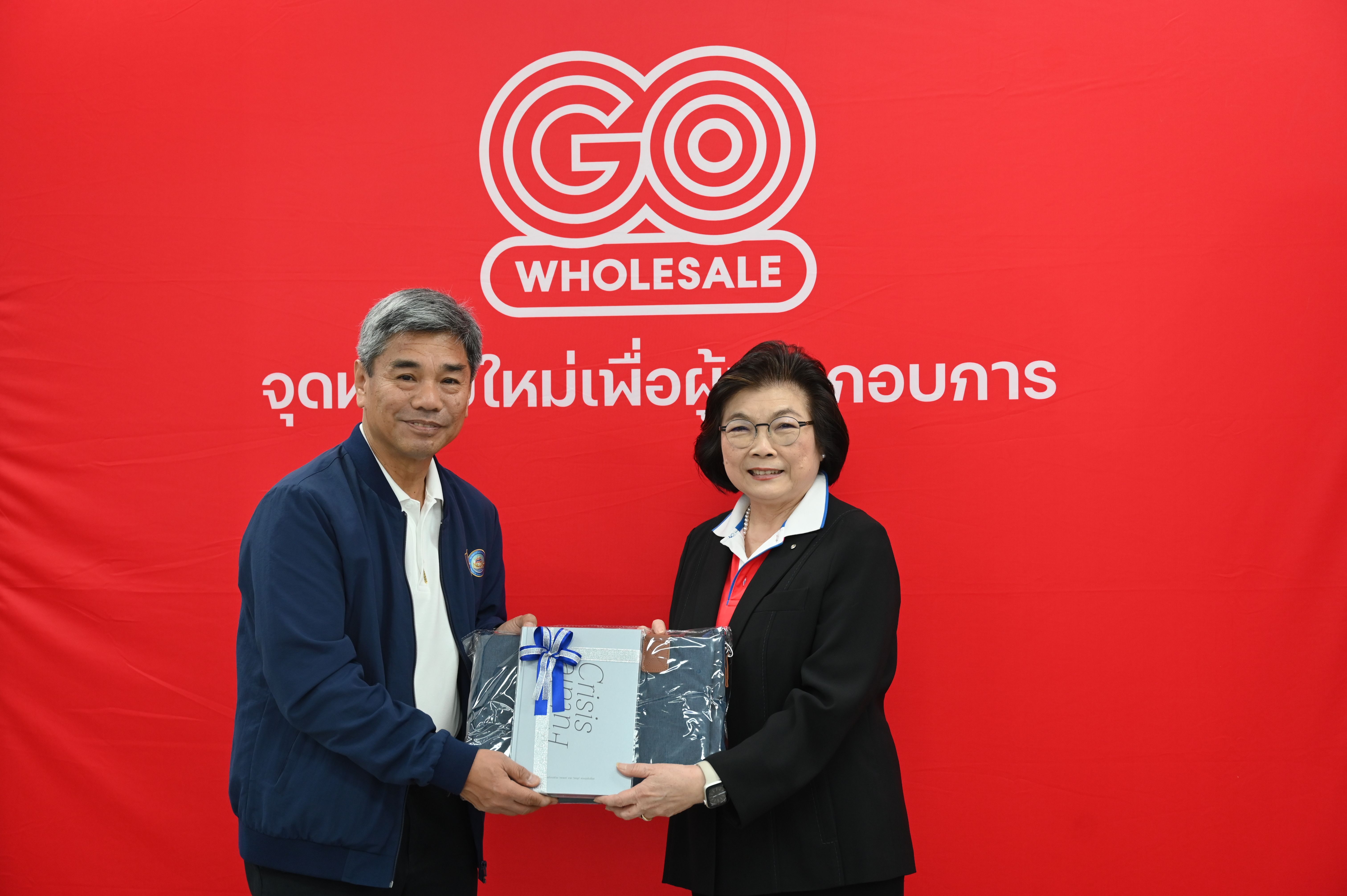Two people exchange a silver gift in front of a red GO WHOLESALE wall.