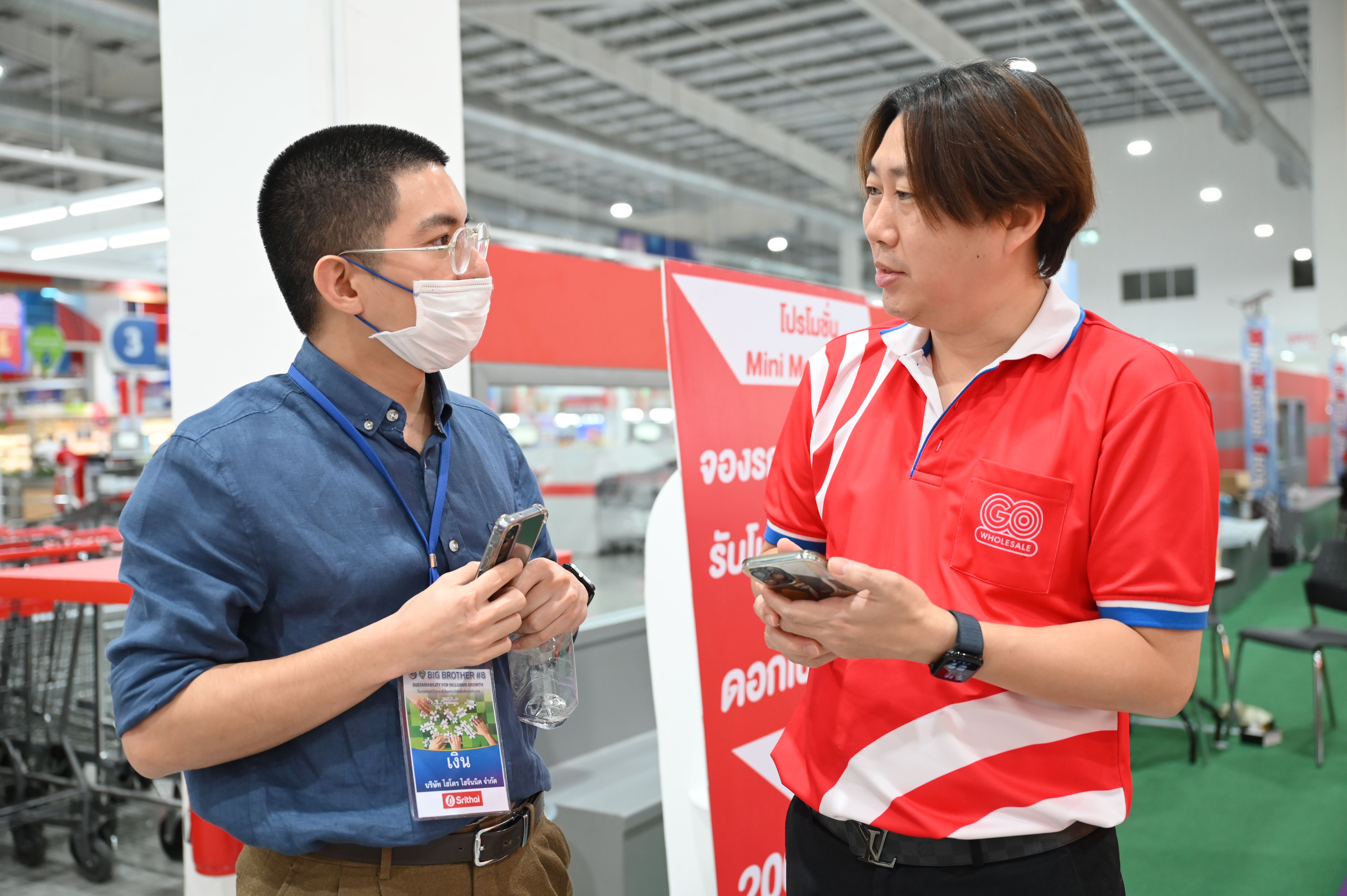 Two men, one masked, one in red polo, conversing in a retail store setting.