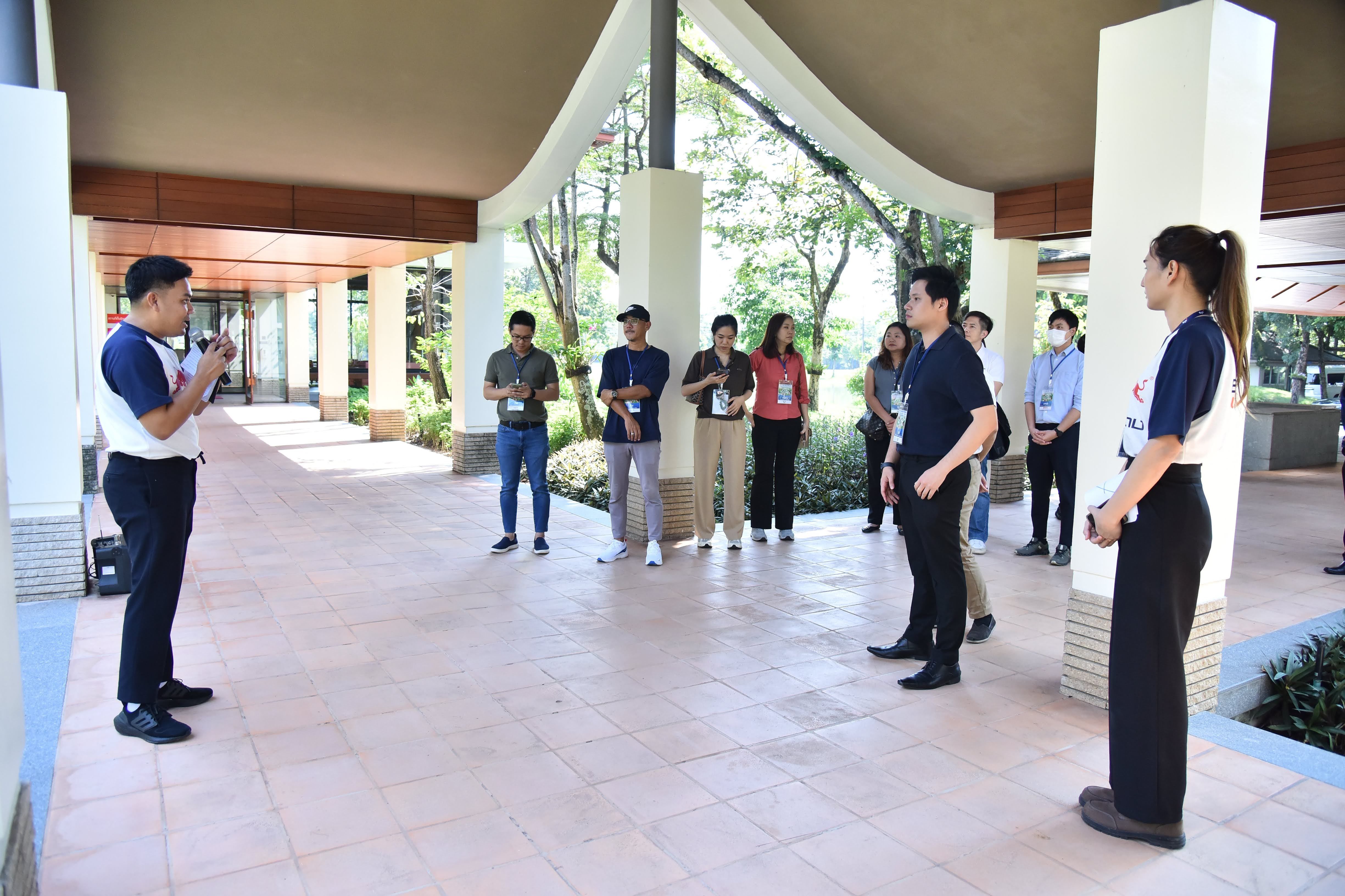 A man speaks to a woman while a group observes in a modern open-air hall.
