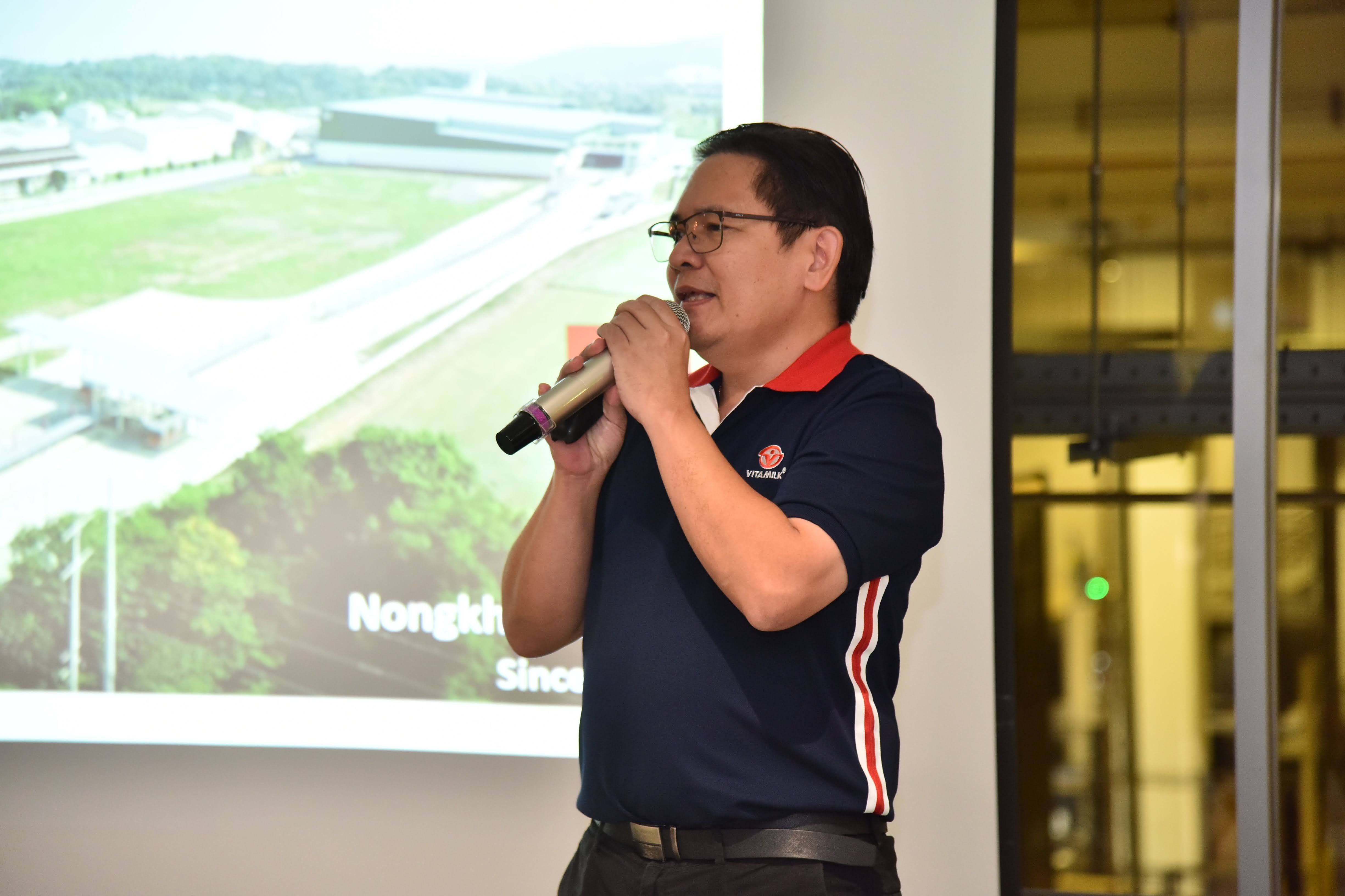 Man in polo shirt speaking into microphone before projector showing aerial landscape.