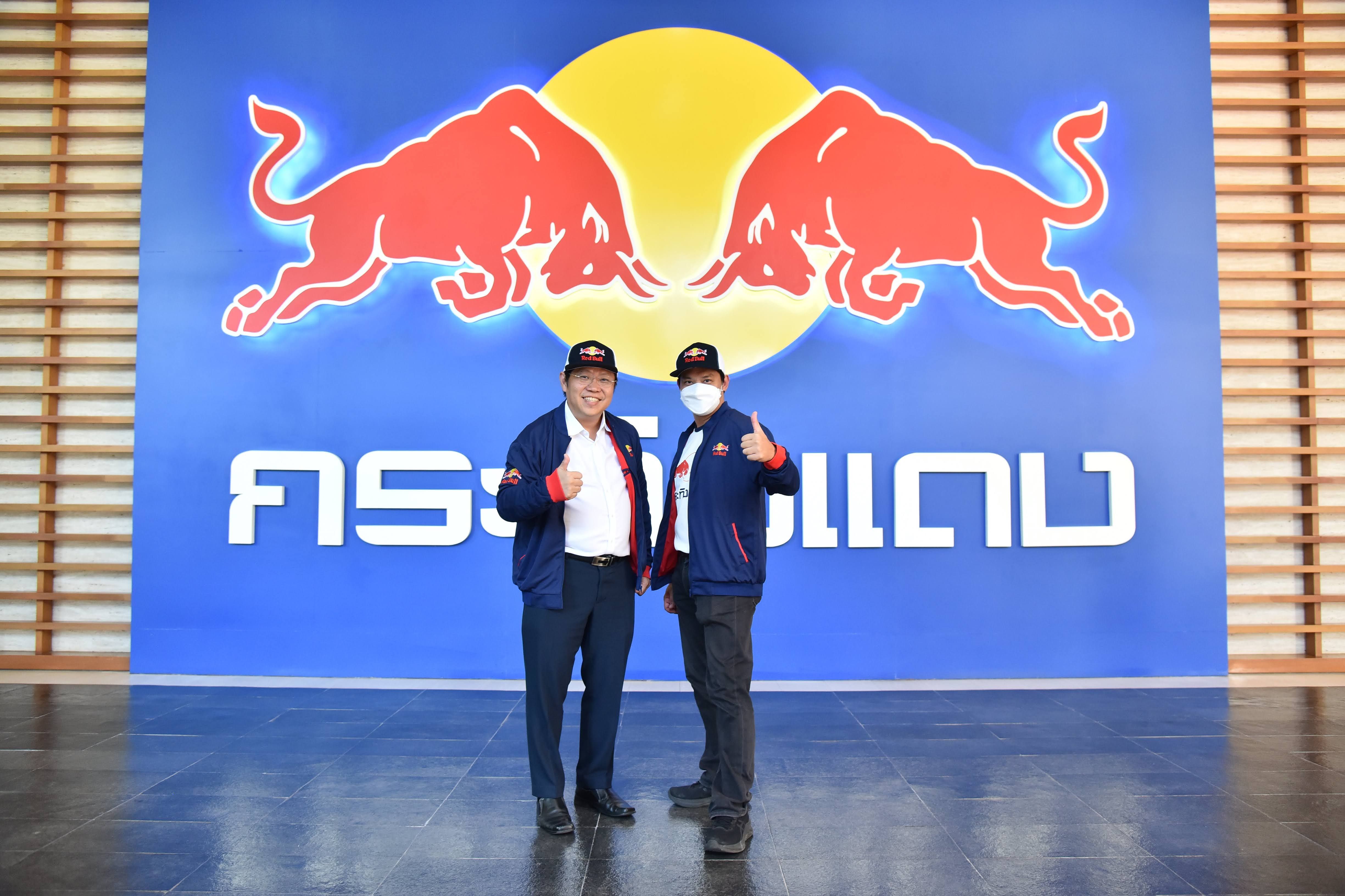 Two men in caps give thumbs-up before a large Red Bull logo with Thai text.