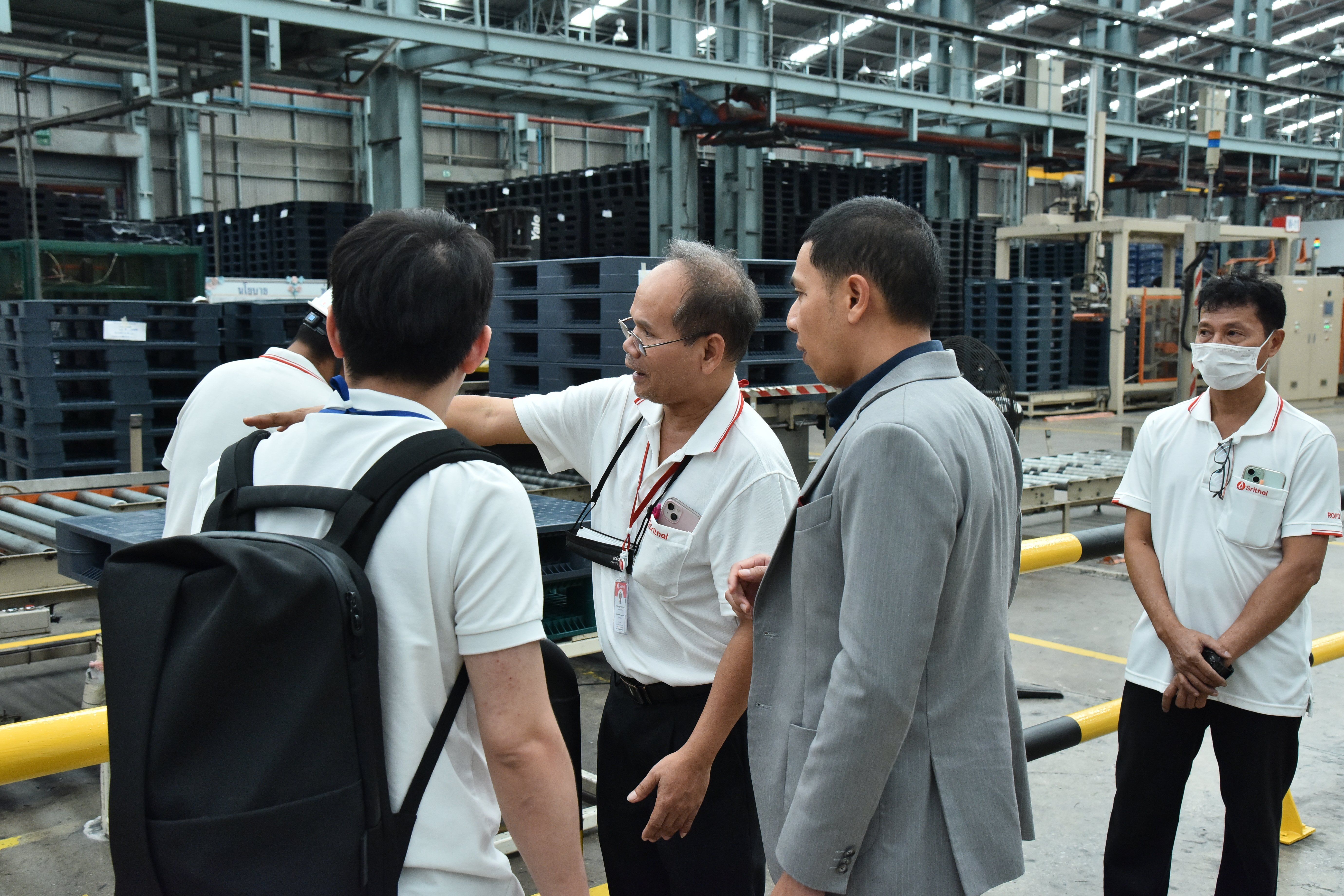 Men in a factory discuss operations, surrounded by industrial equipment and pallets.