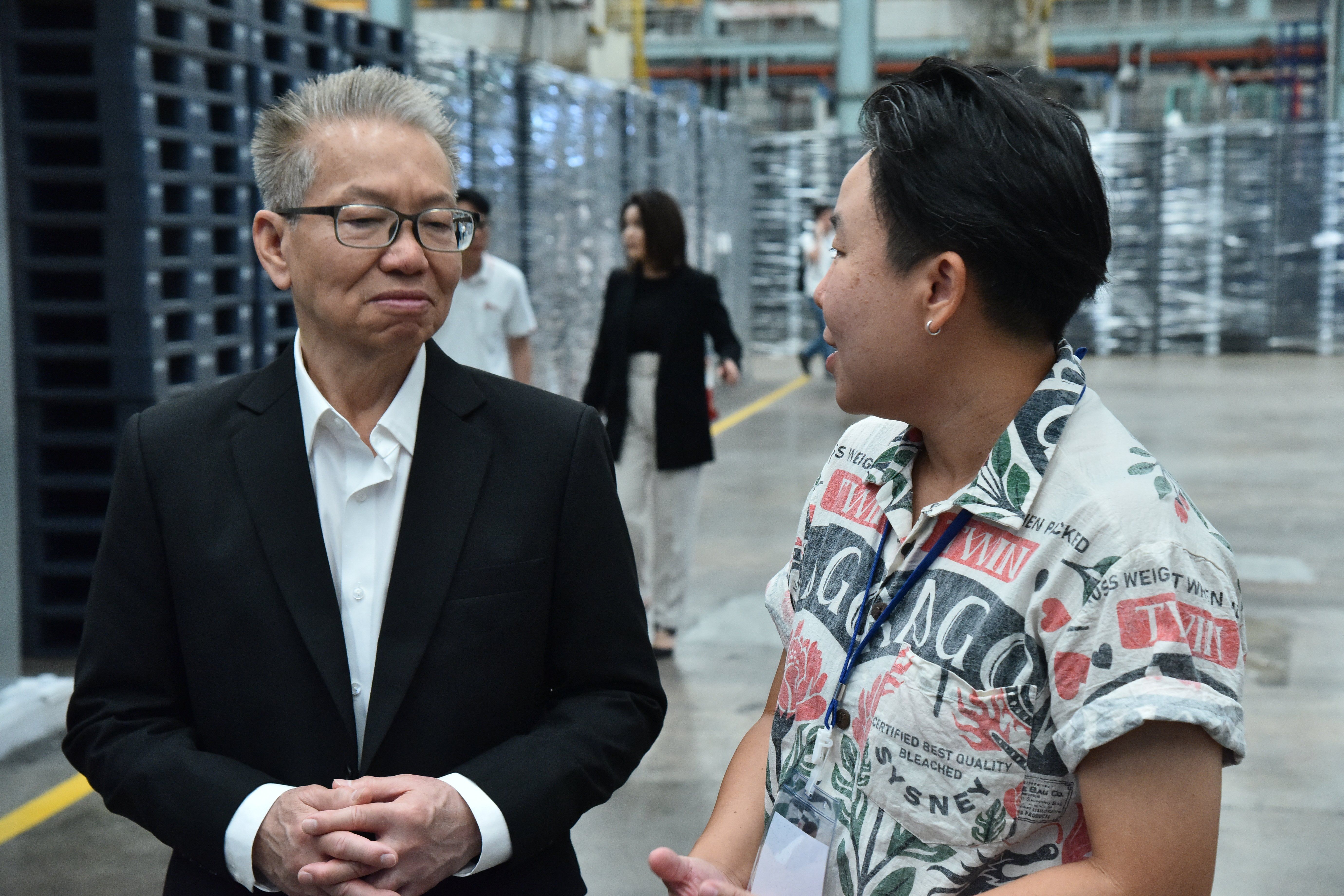 Two men, one in a suit and one in a patterned shirt, conversing in a warehouse.