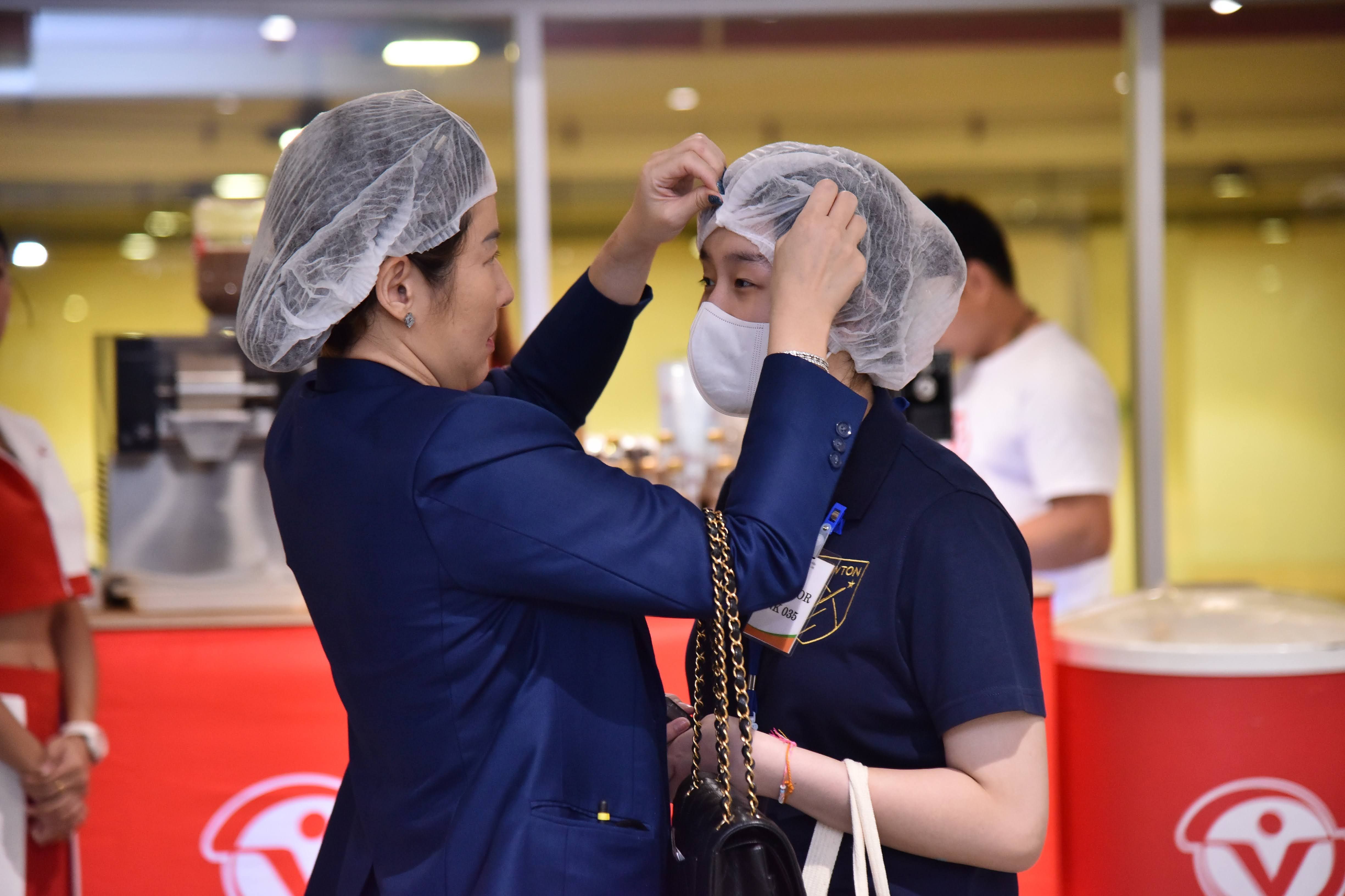 A person helps another put on a hairnet and mask in a clean environment.