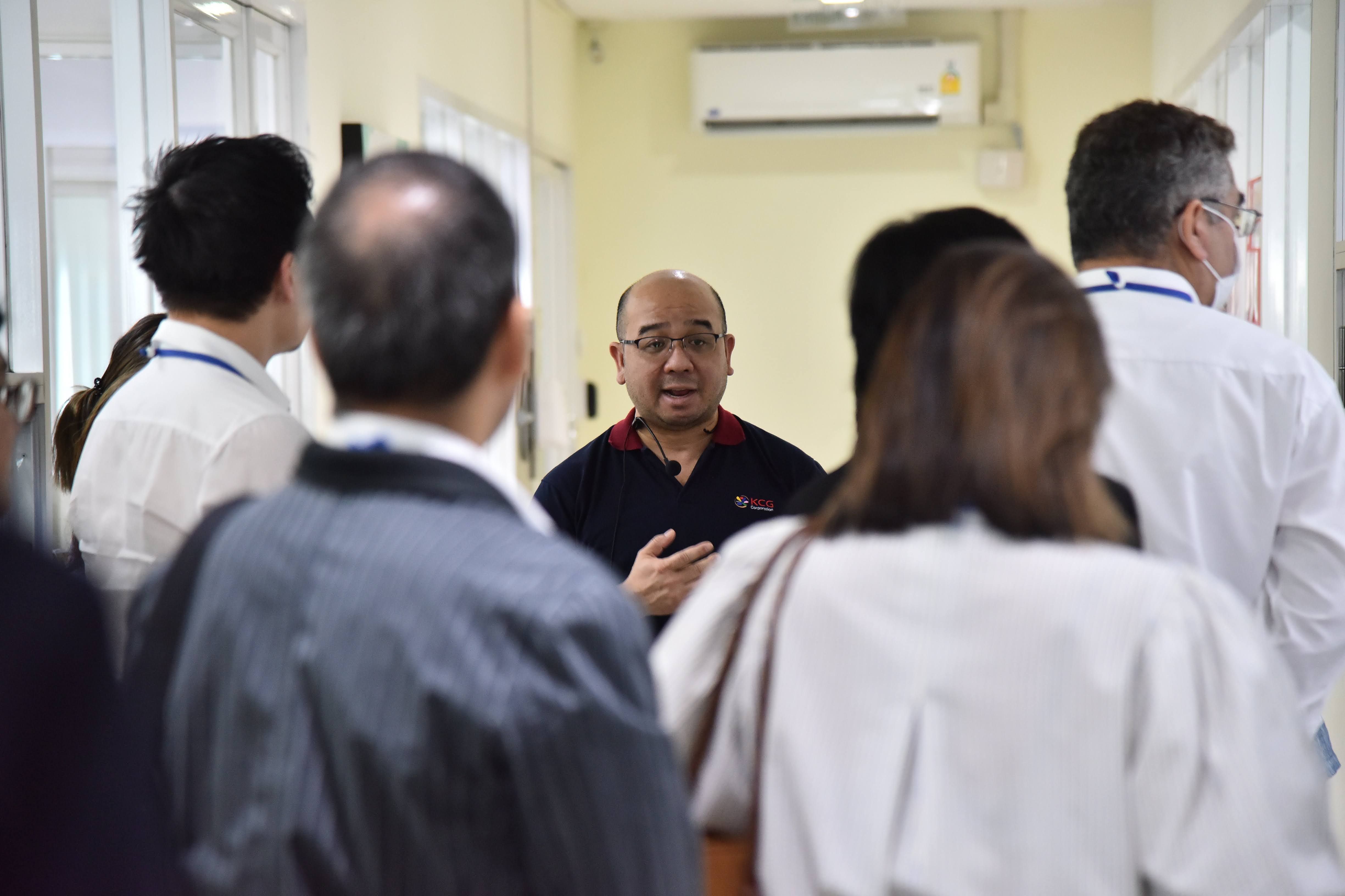 Man in dark shirt and glasses speaking to an attentive group of people indoors.