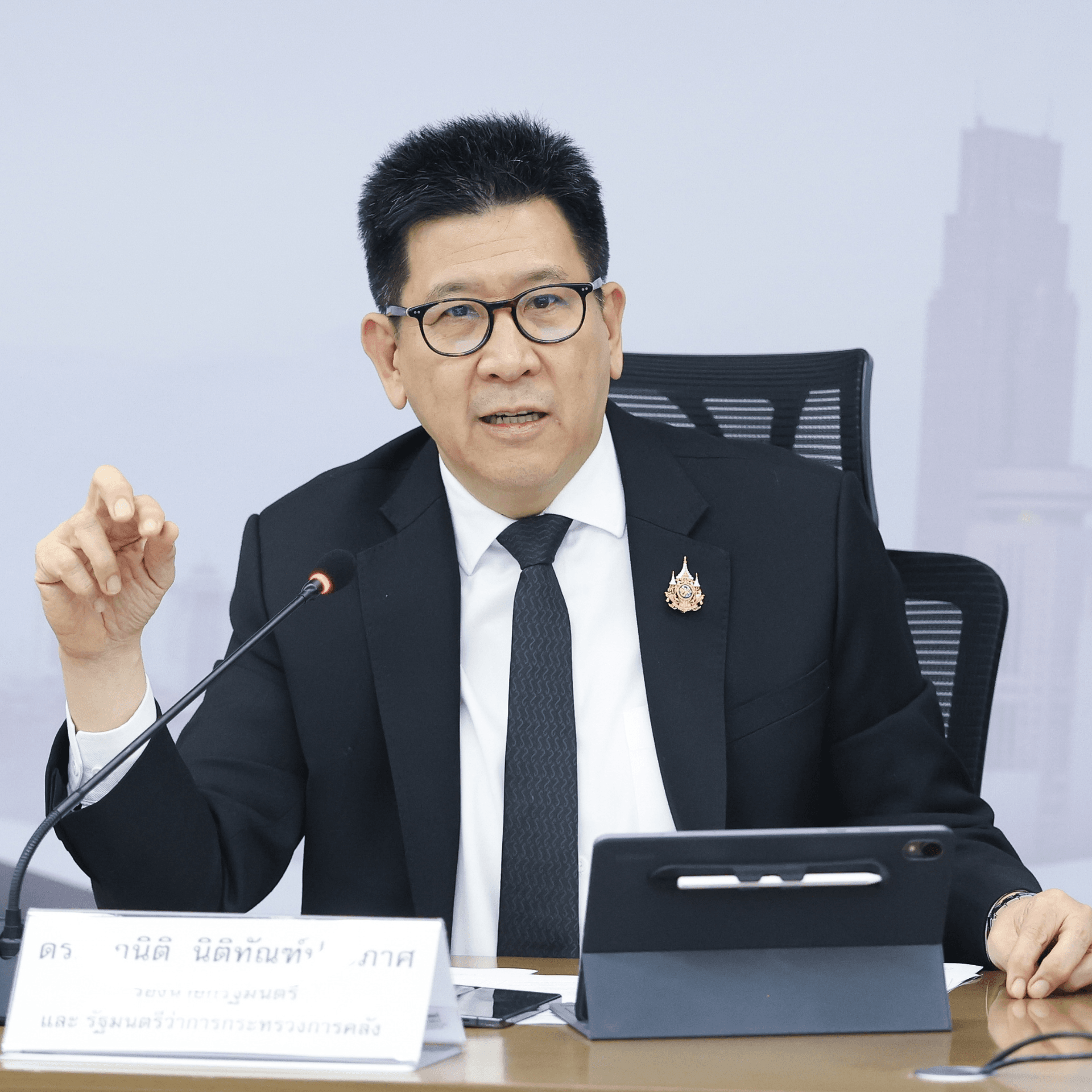 Professional man in dark suit and glasses speaks, gesturing at a conference table.