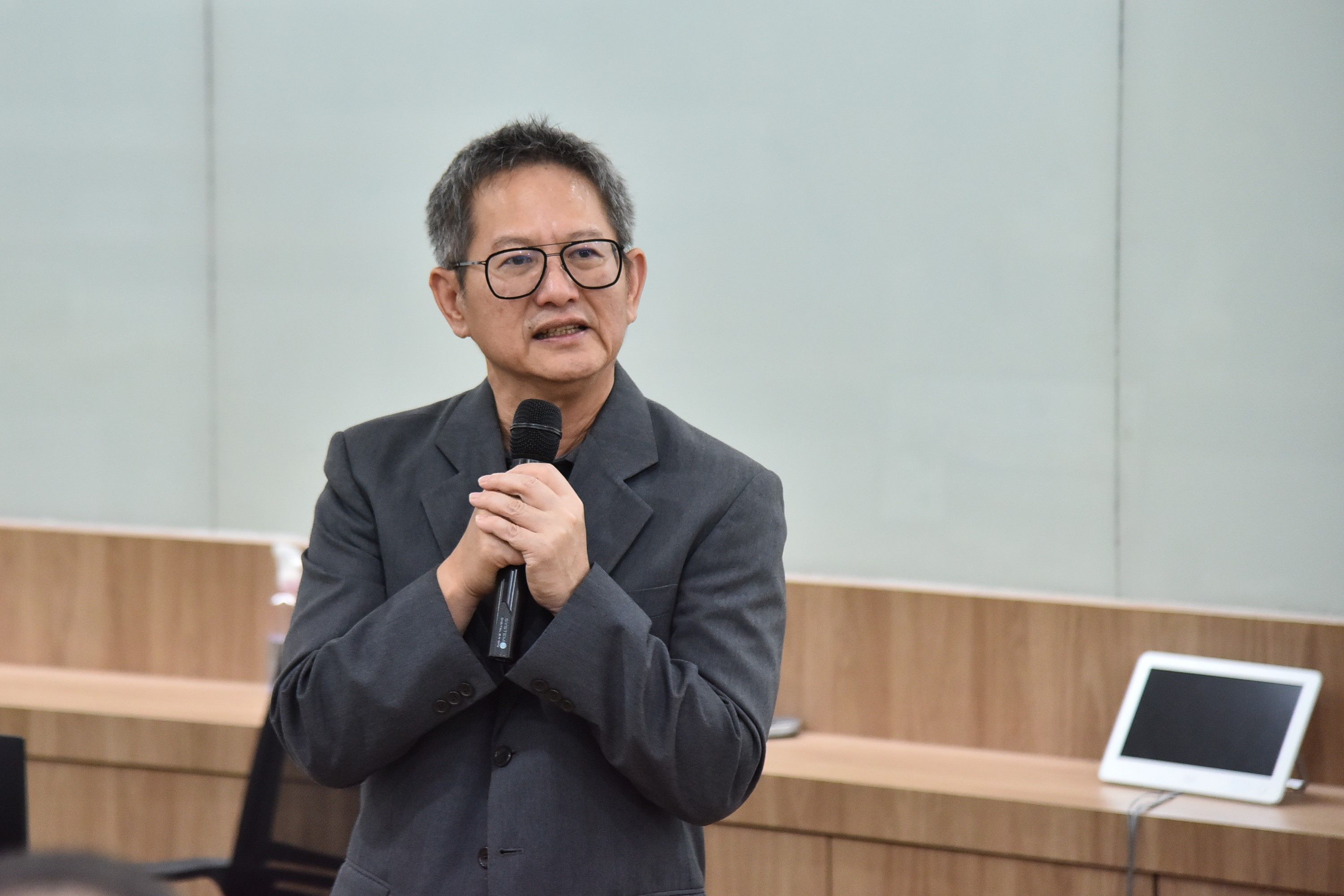 Grey-haired man in glasses speaks into a microphone, delivering a presentation.
