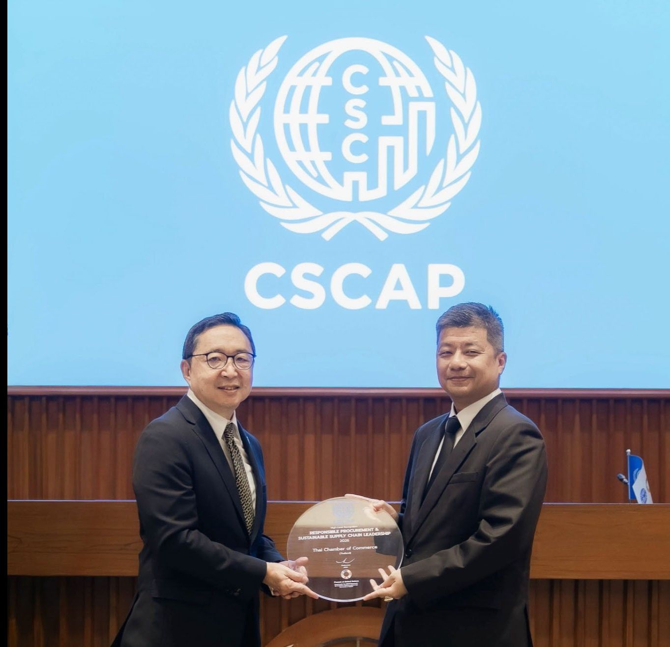 Two men in suits exchange a spherical award against a blue wall with CSCAP and UN logos.