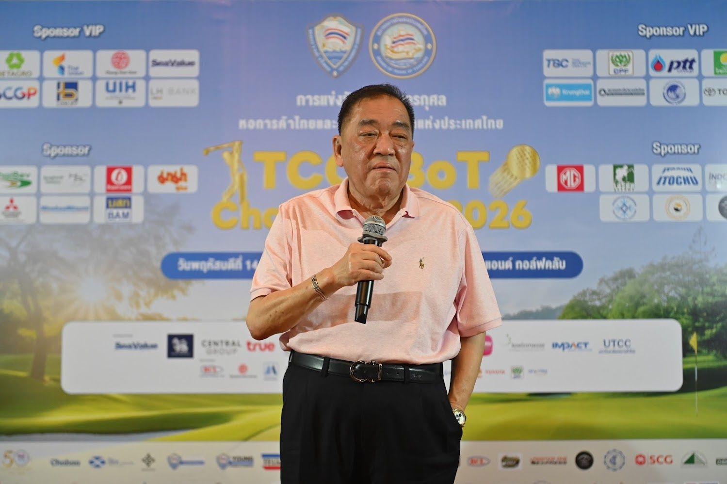 Man in pink polo shirt speaks into microphone at a sponsored event.