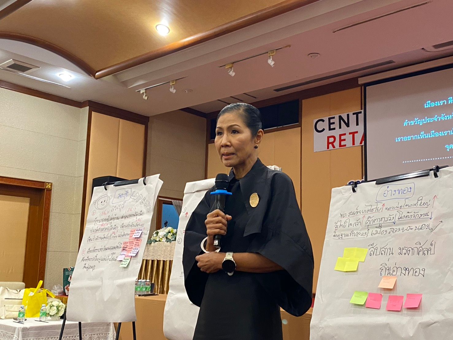 Woman in black dress speaks into microphone at a conference with flip charts.