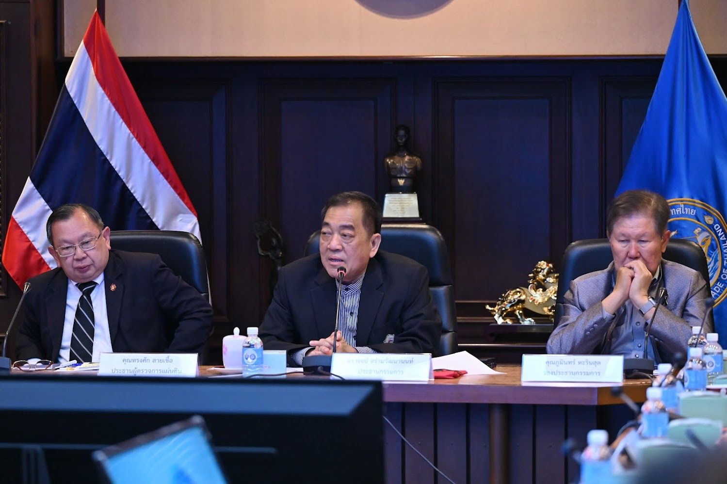 Three men in suits seated at a meeting table with a Thai flag in the background.