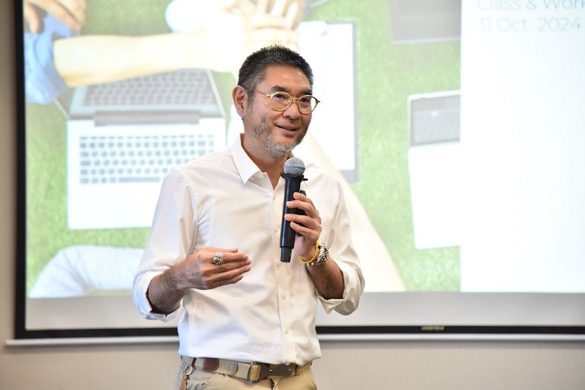 Man in glasses and white shirt speaks into microphone, gesturing during presentation.