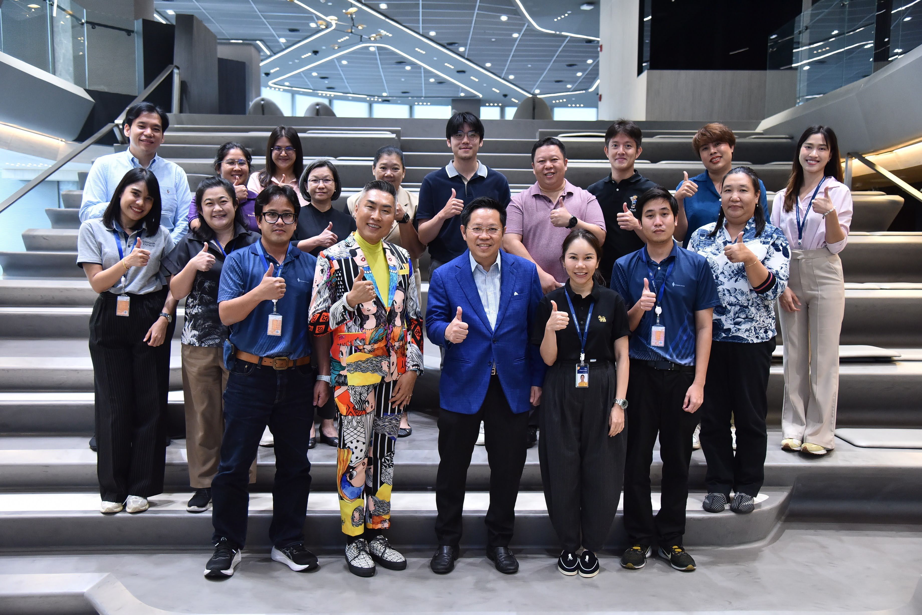 A large group of smiling people giving thumbs-up on a modern indoor staircase.