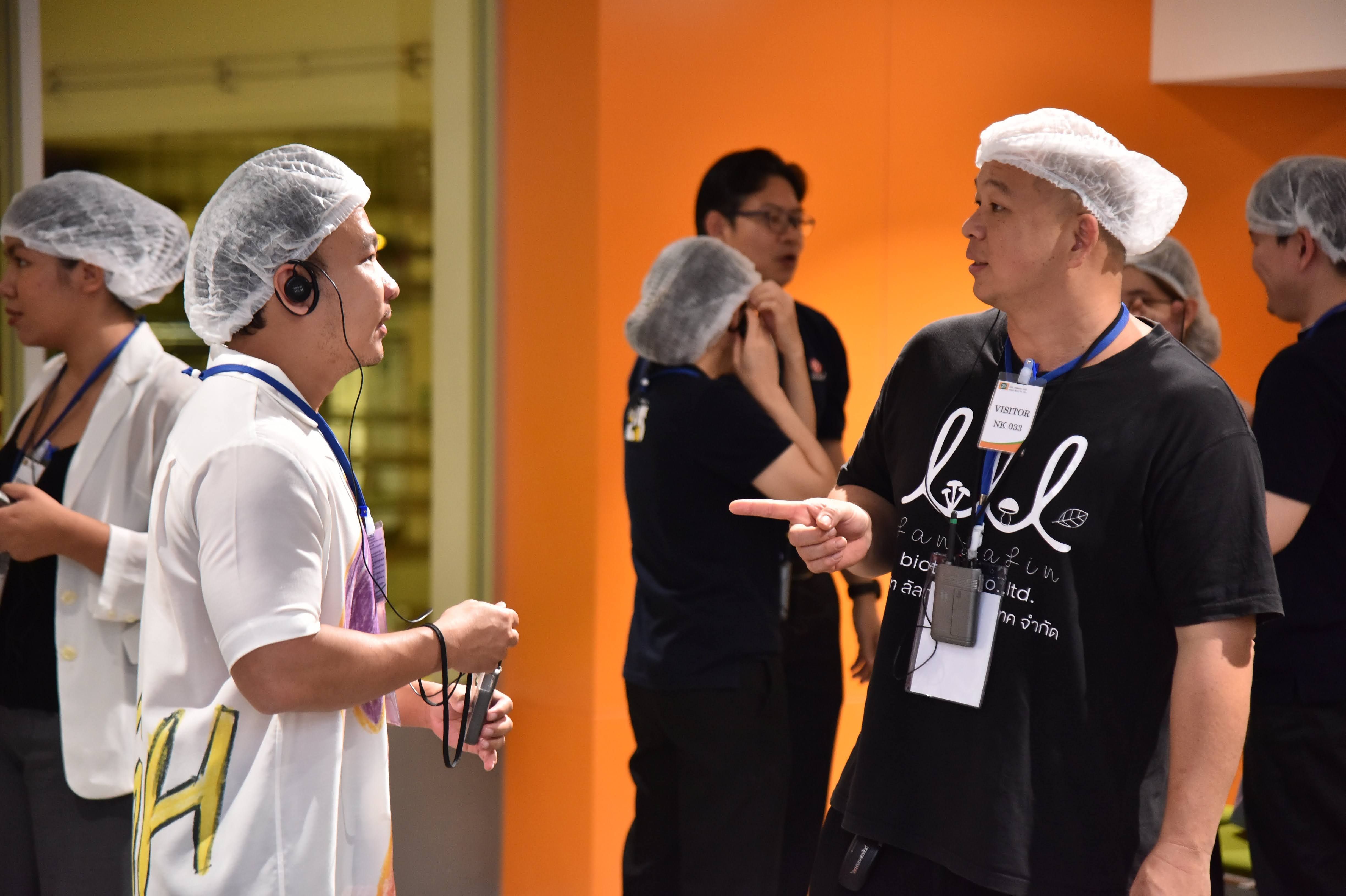 Two men in hairnets converse in an industrial setting, one pointing emphatically.