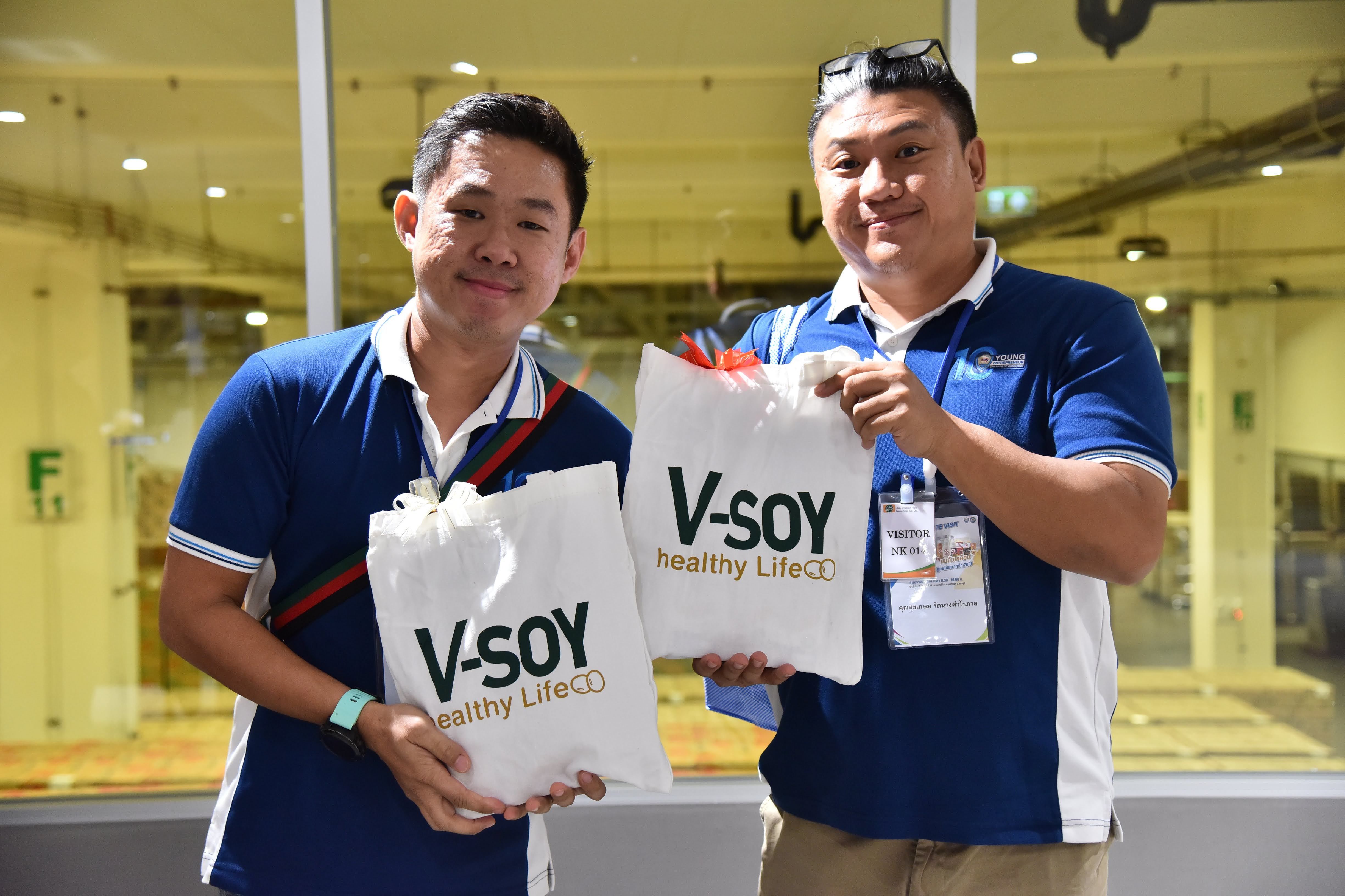 Two smiling men hold white 'V-SOY Healthy Life CO' bags at an indoor event.