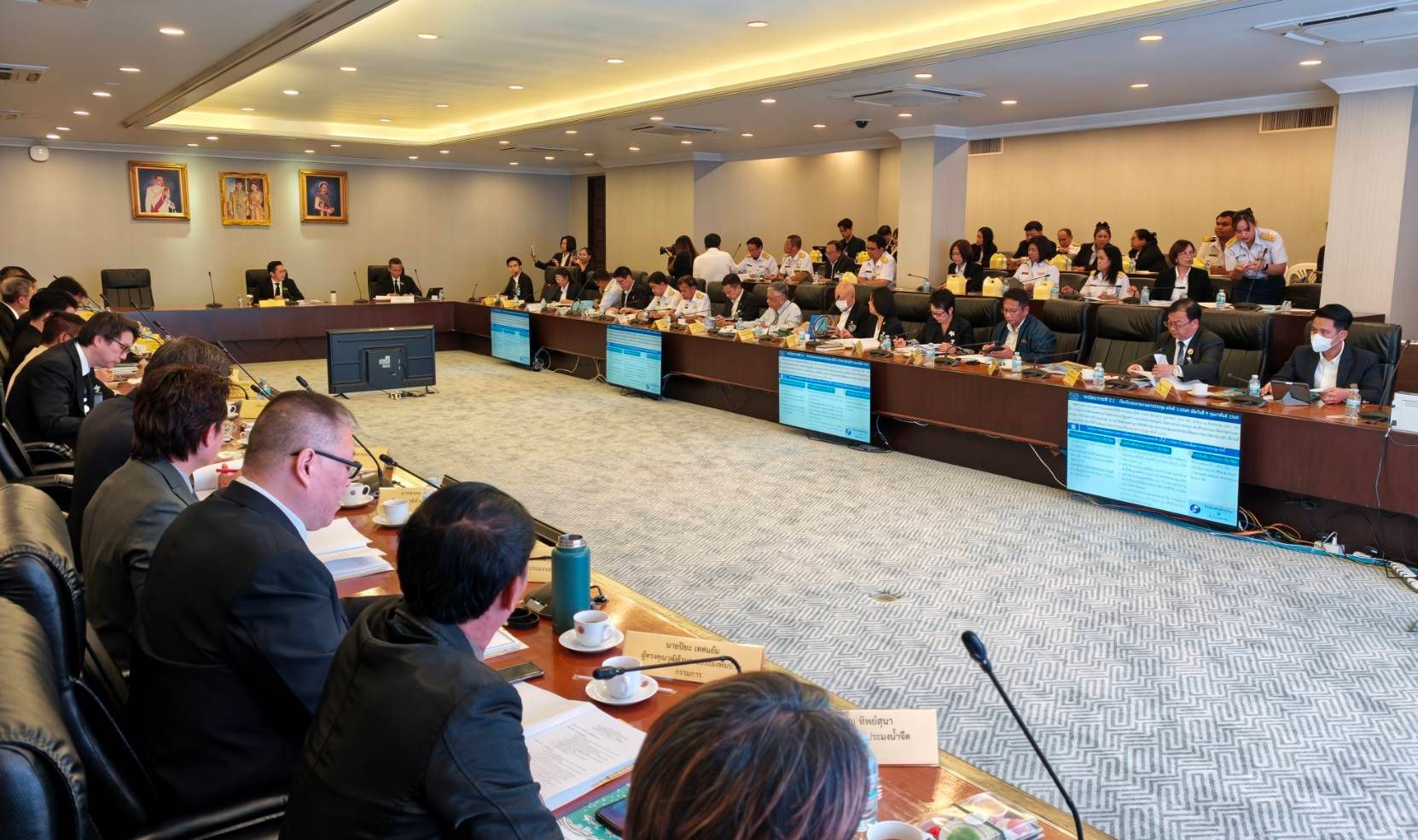 Attendees in a large conference room with multiple screens embedded in a U-shaped table.