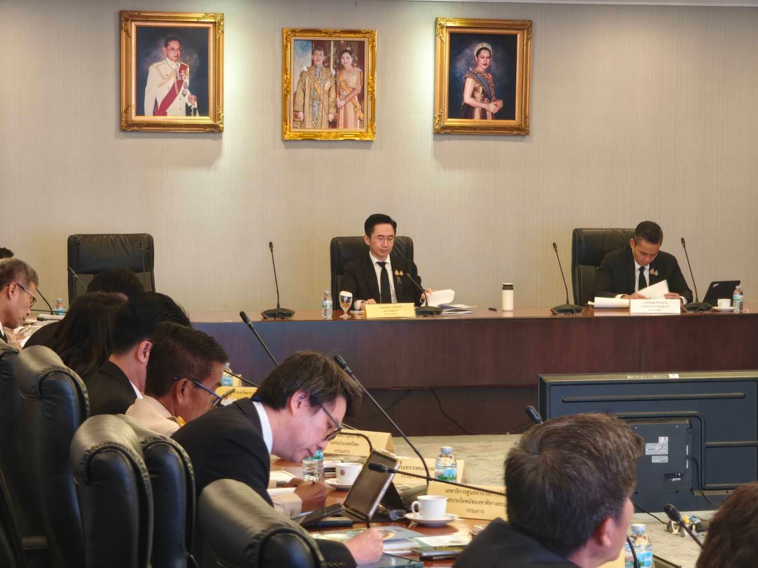 Formal meeting with officials seated at a long table under three framed portraits.