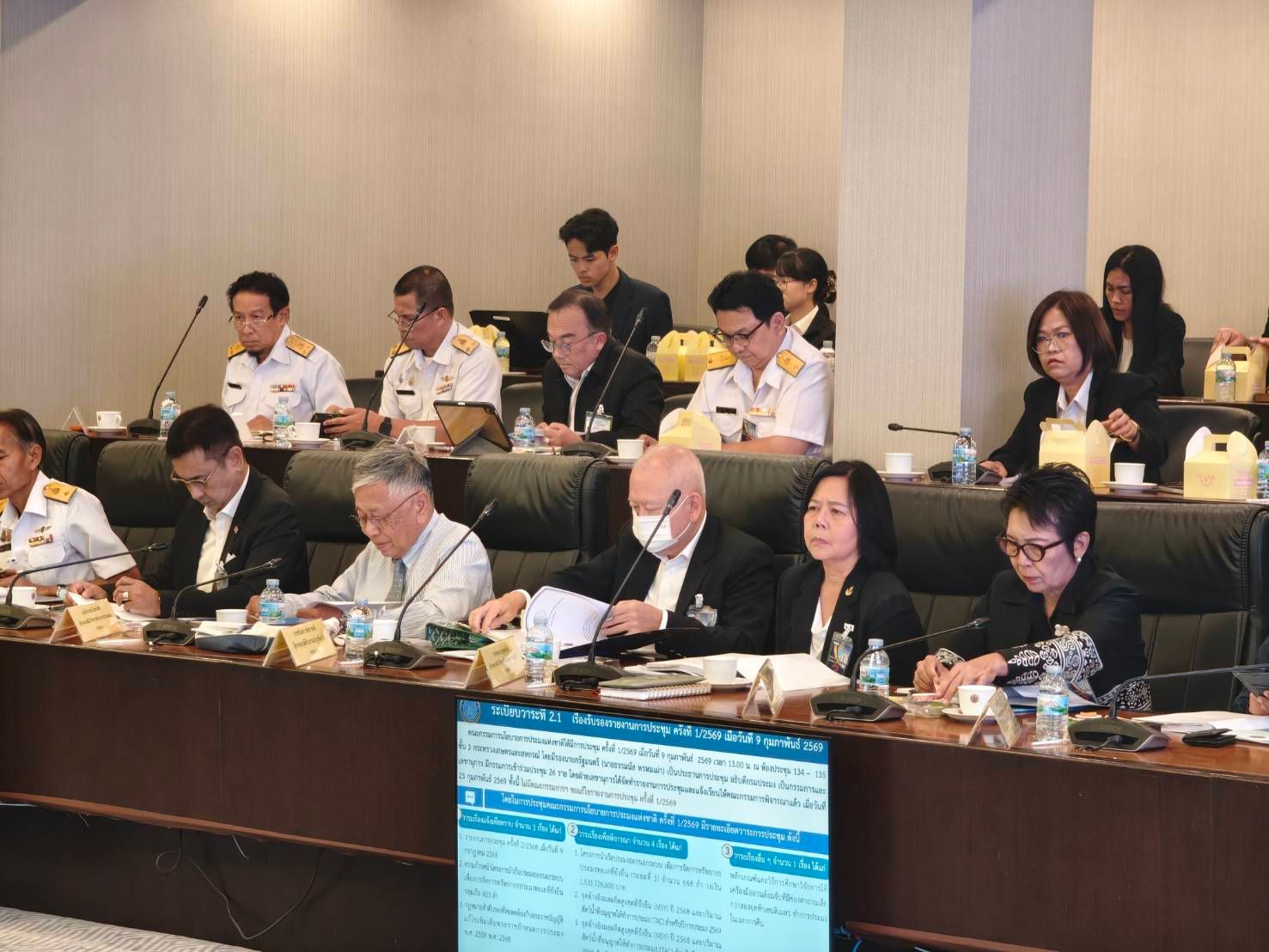 Officials attend a formal meeting around a large conference table with a screen display.