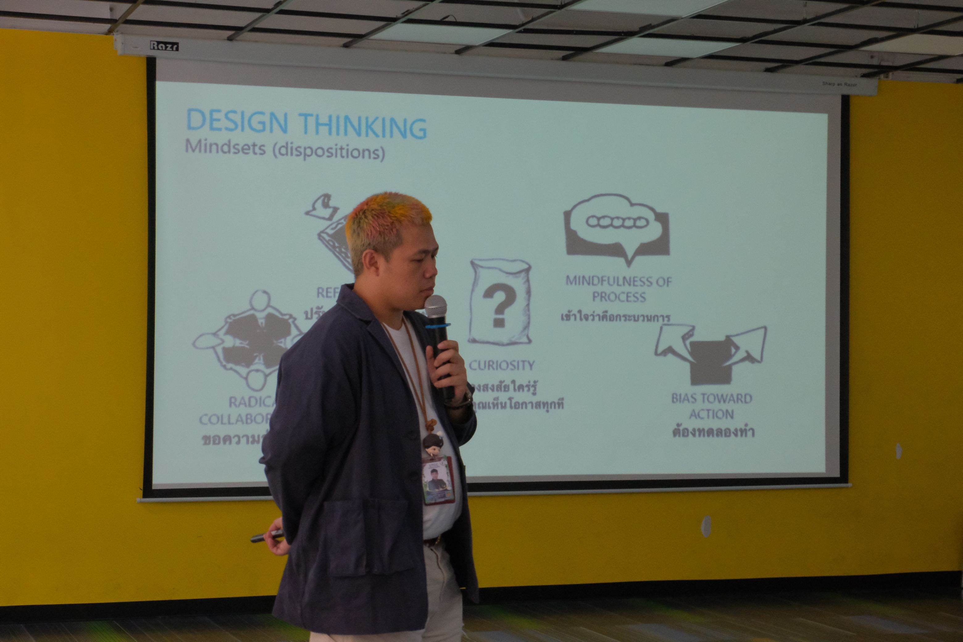 Man speaks into microphone in front of a 'Design Thinking' presentation screen.