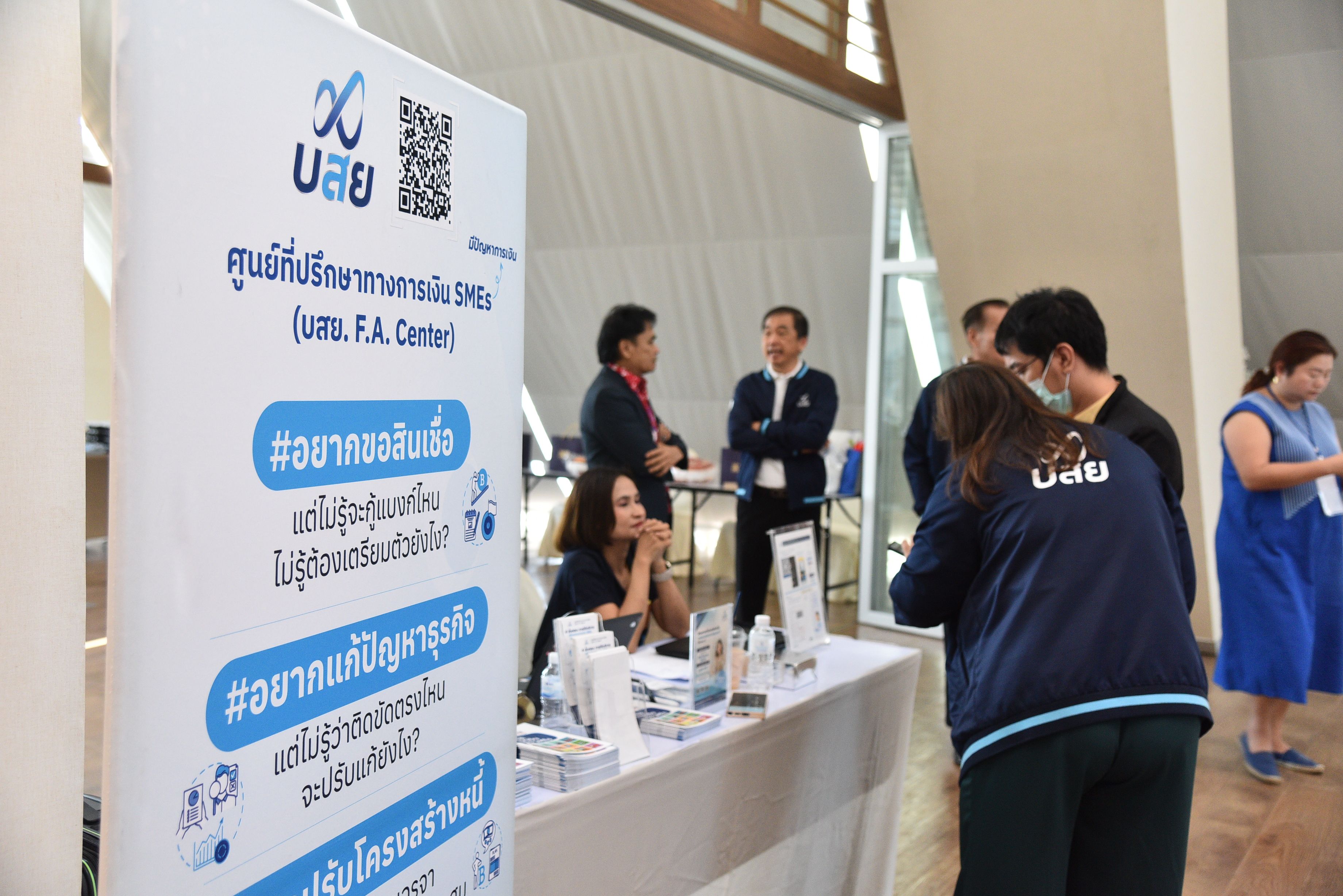 People interact near a blue and white promotional banner displaying Thai text at an indoor event.