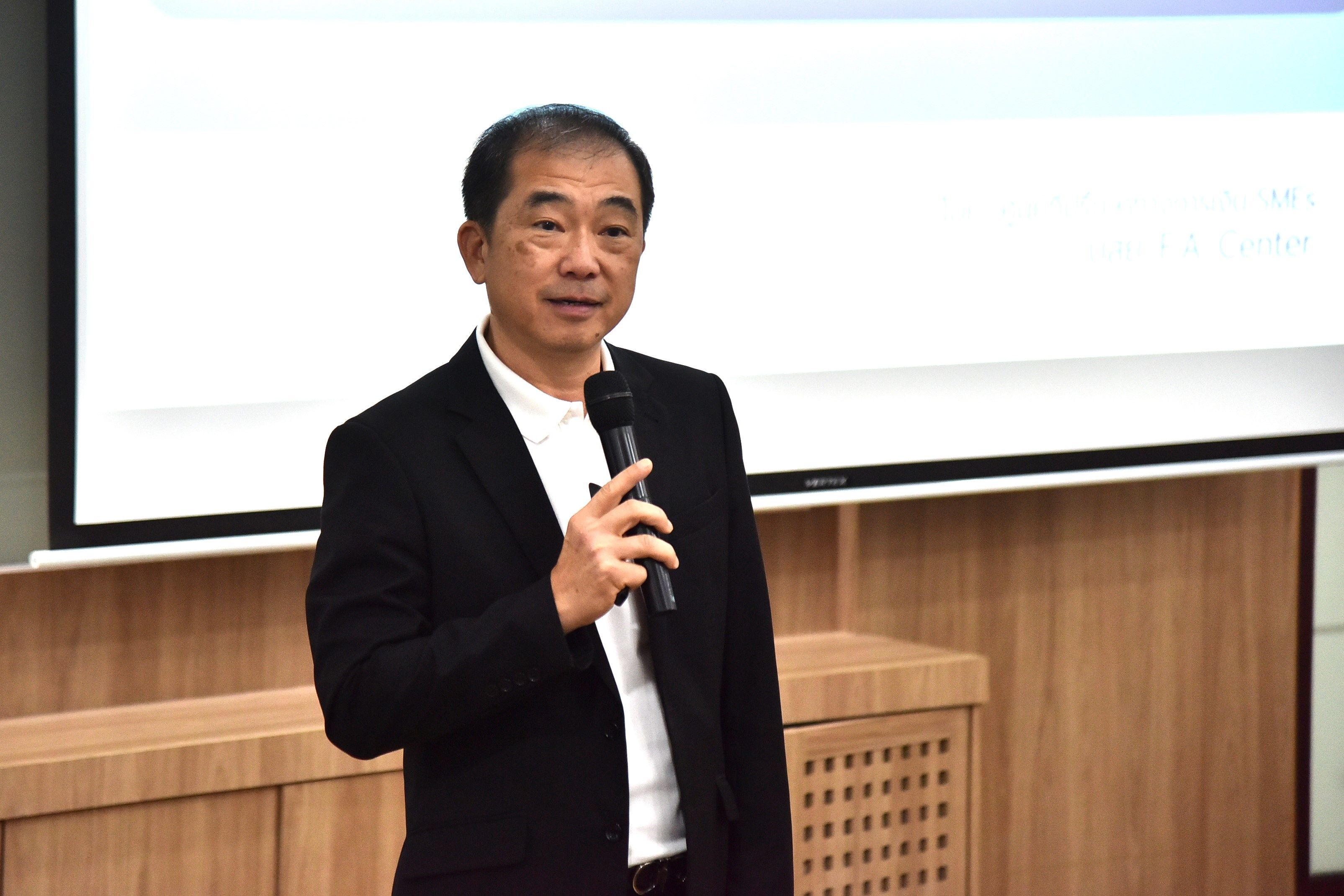 An Asian man in a black jacket speaks into a microphone during a presentation.