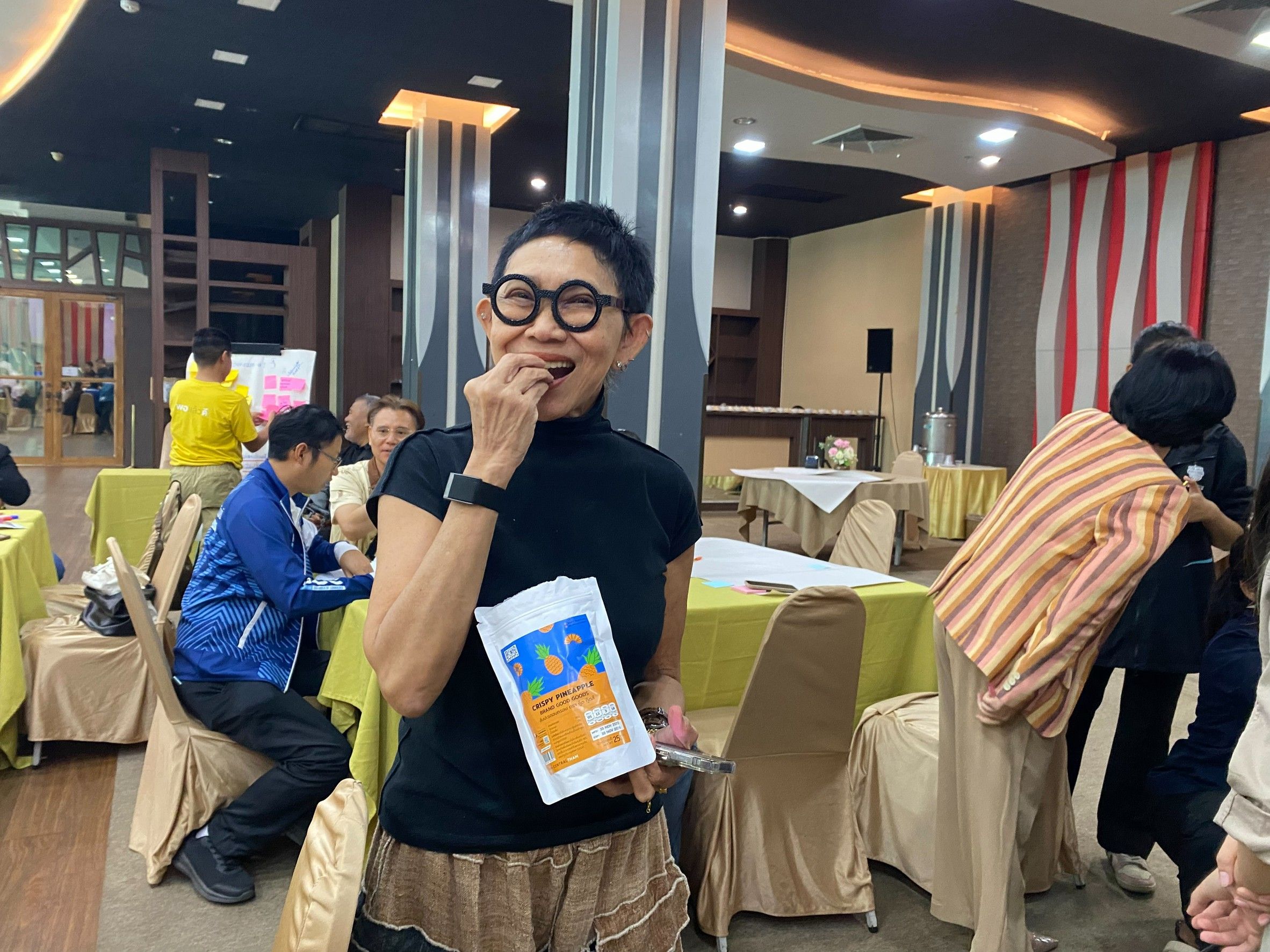 Woman with glasses smiles eating a snack from a packet in a busy room.