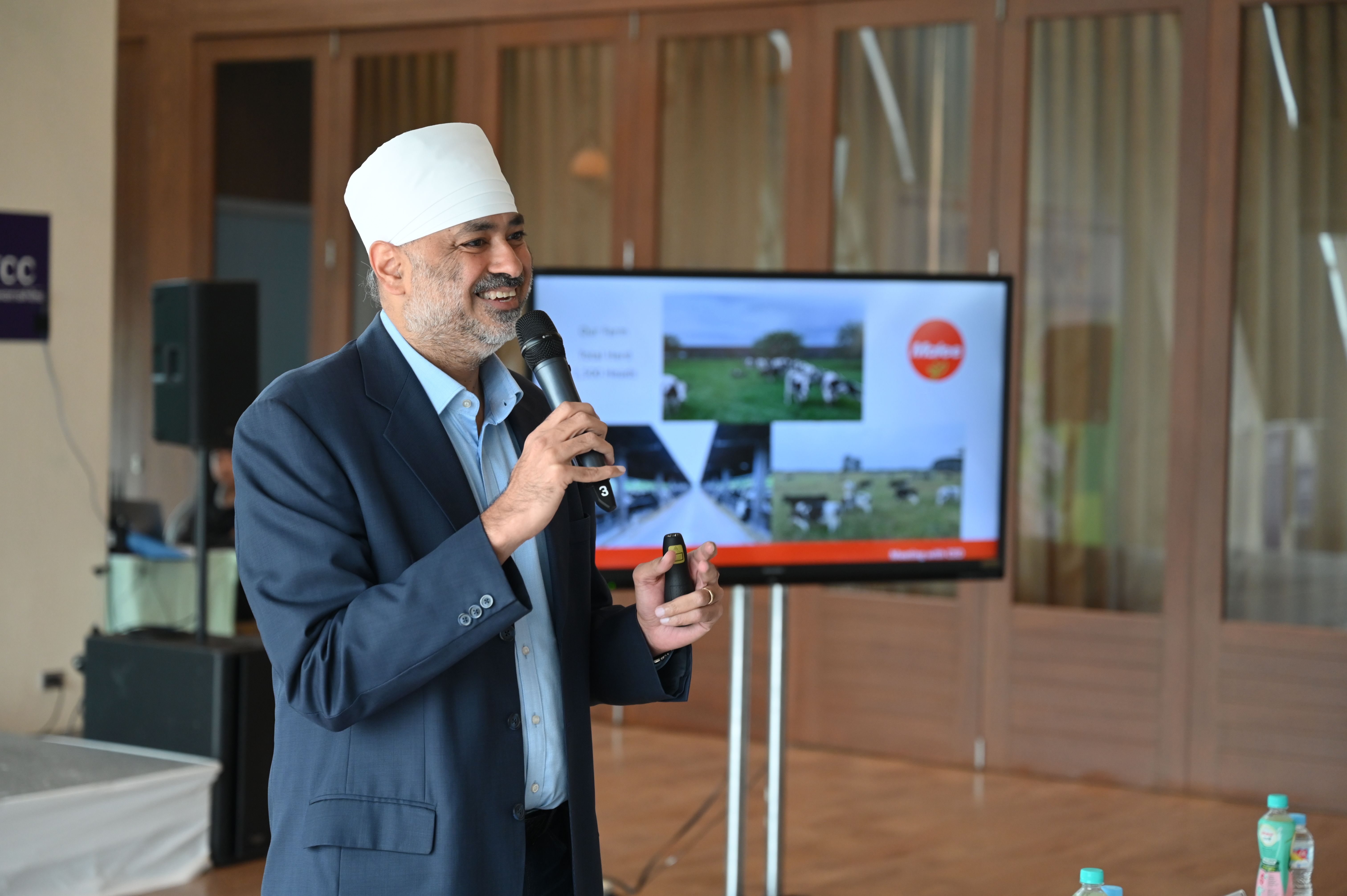 Man in suit and white cap speaks into microphone, presenting images of cows on screen.