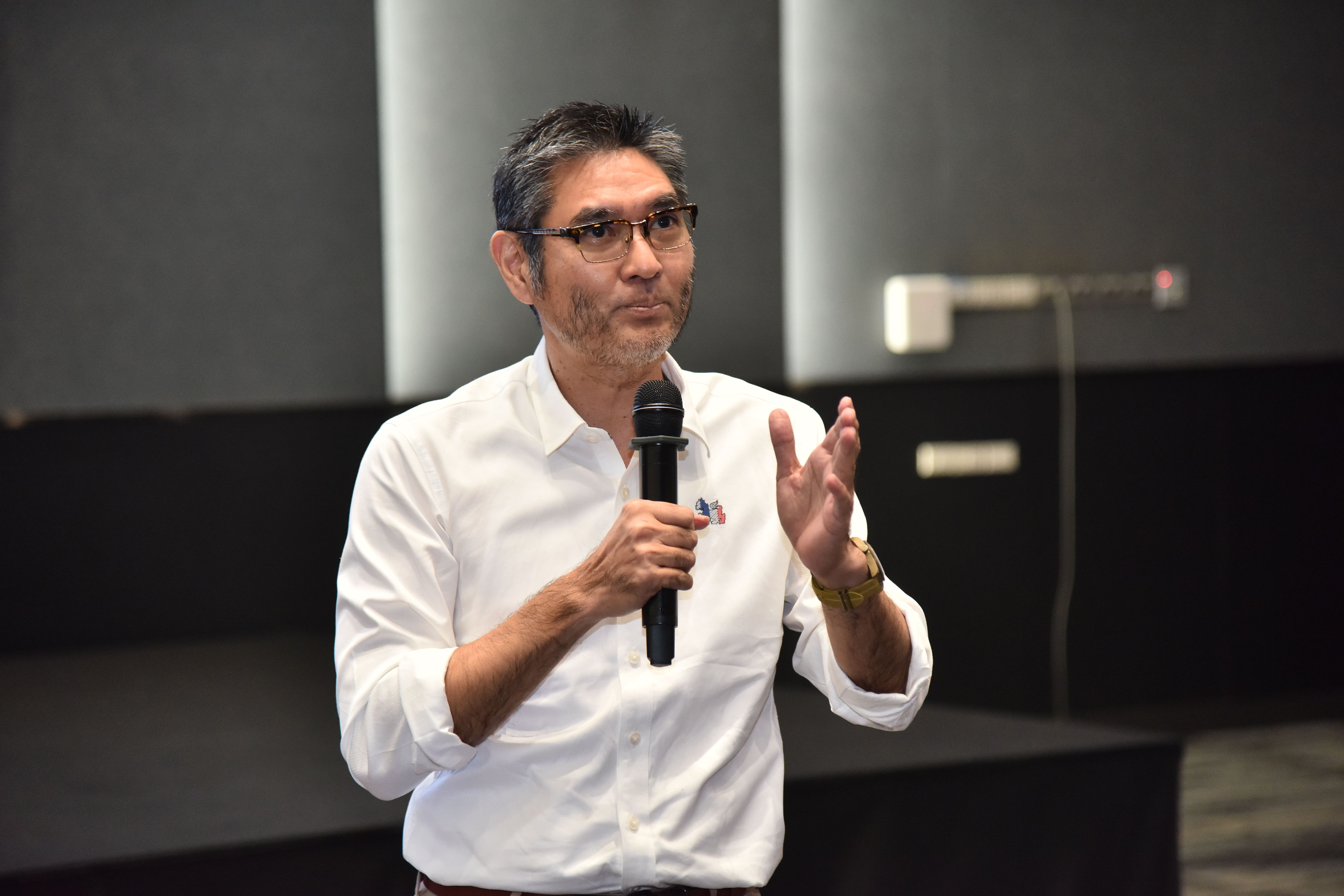Man with glasses speaking into a microphone, gesturing with his hand during a presentation.