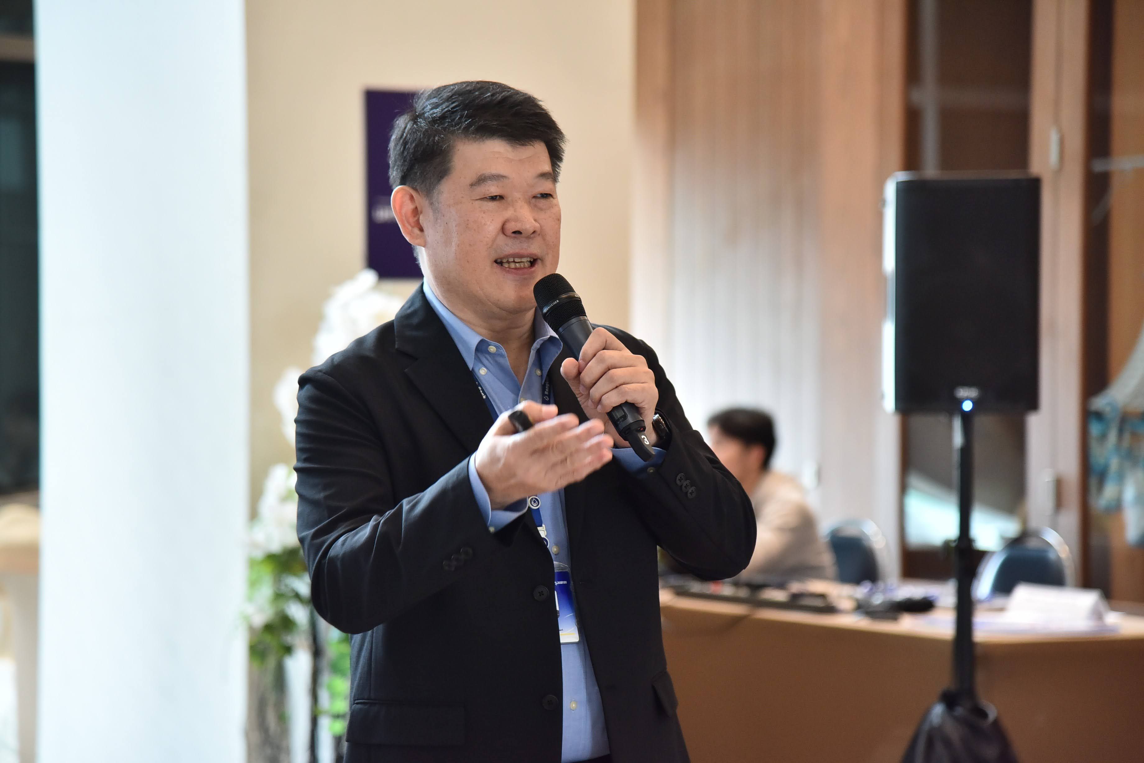 Man in a suit jacket speaks into a microphone, gesturing during an indoor presentation.