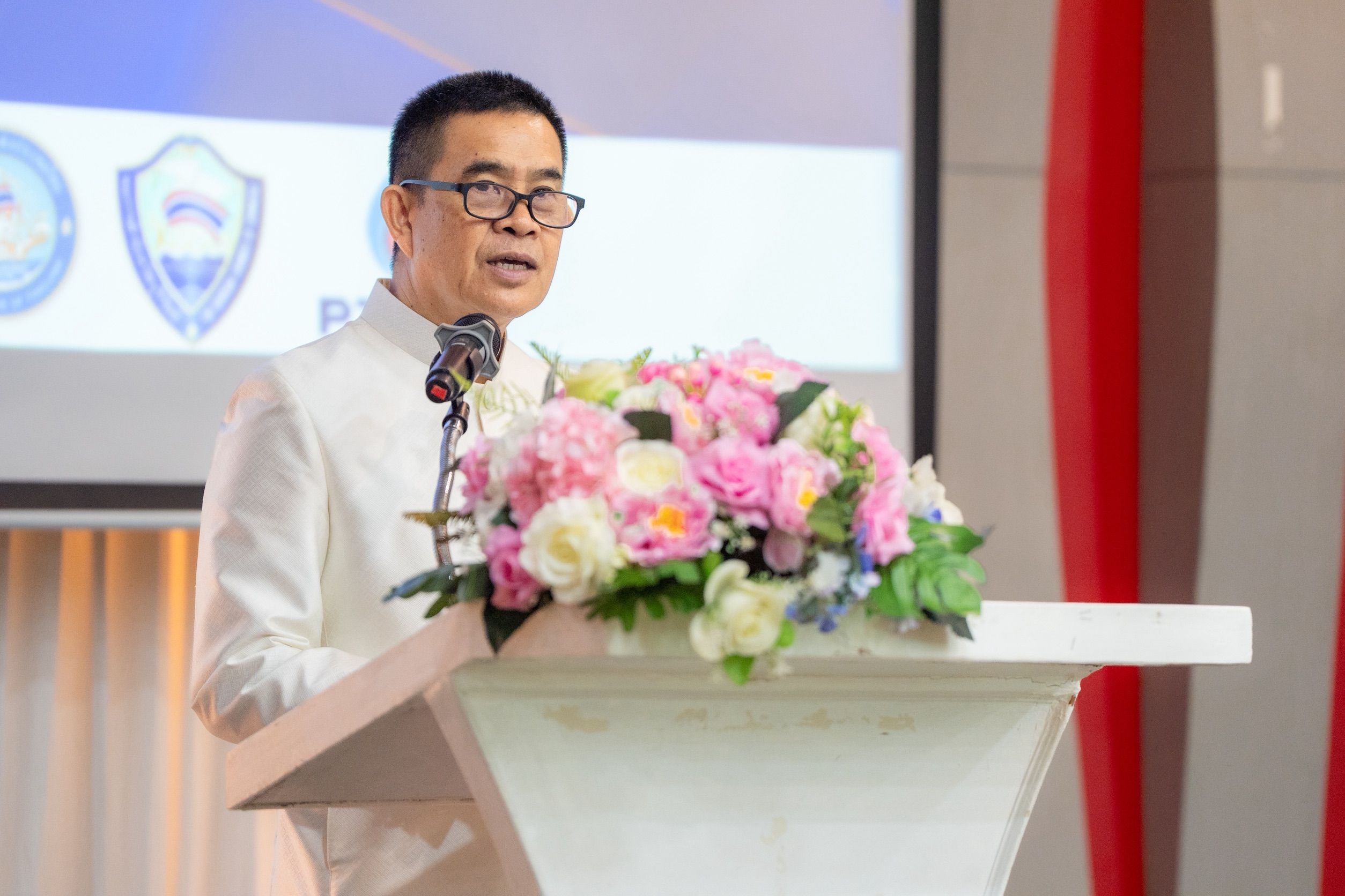 Man in white suit and glasses speaks at a podium with a floral arrangement.