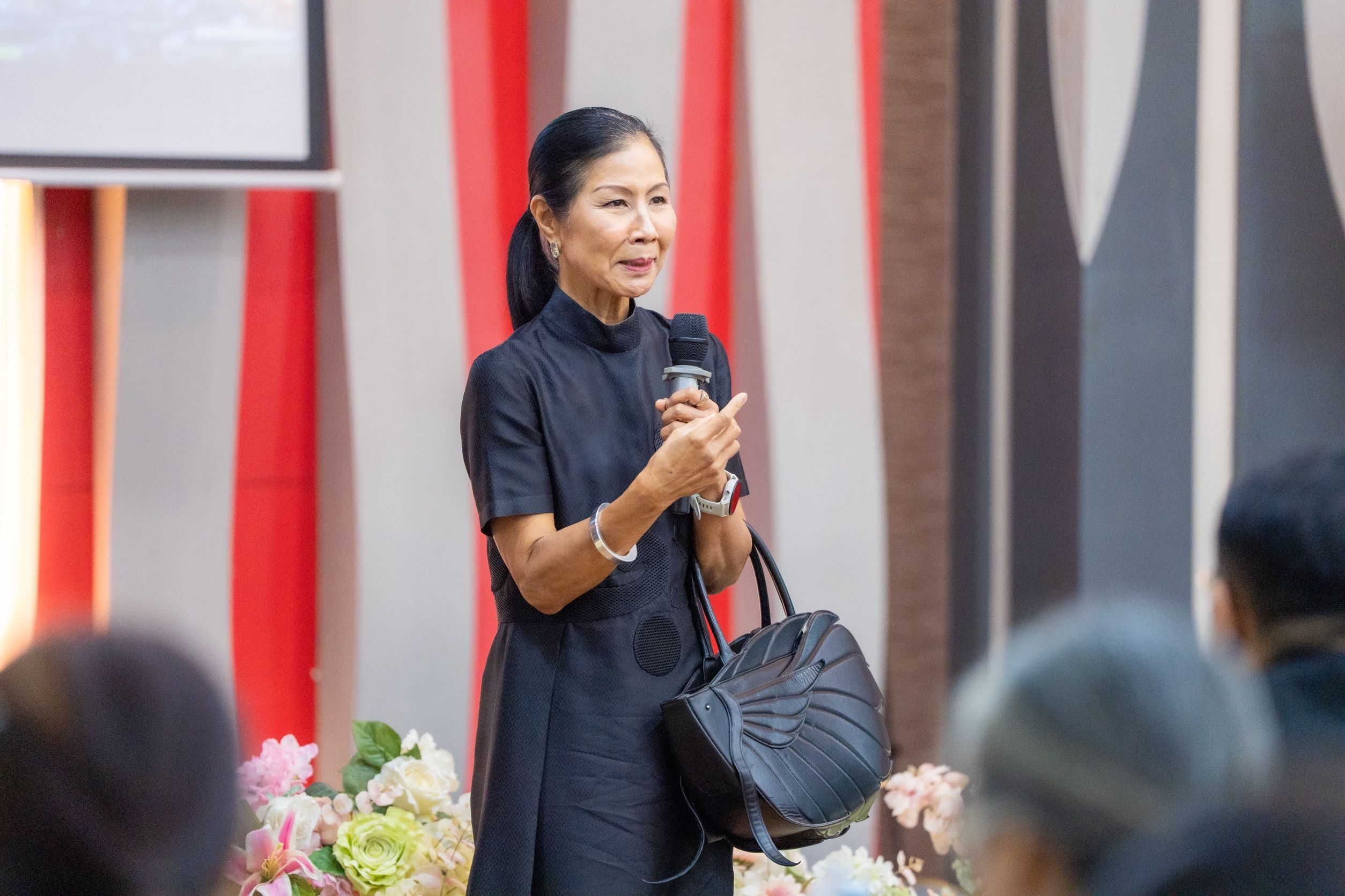 An elegant Asian woman in a black dress speaks into a microphone at an event.