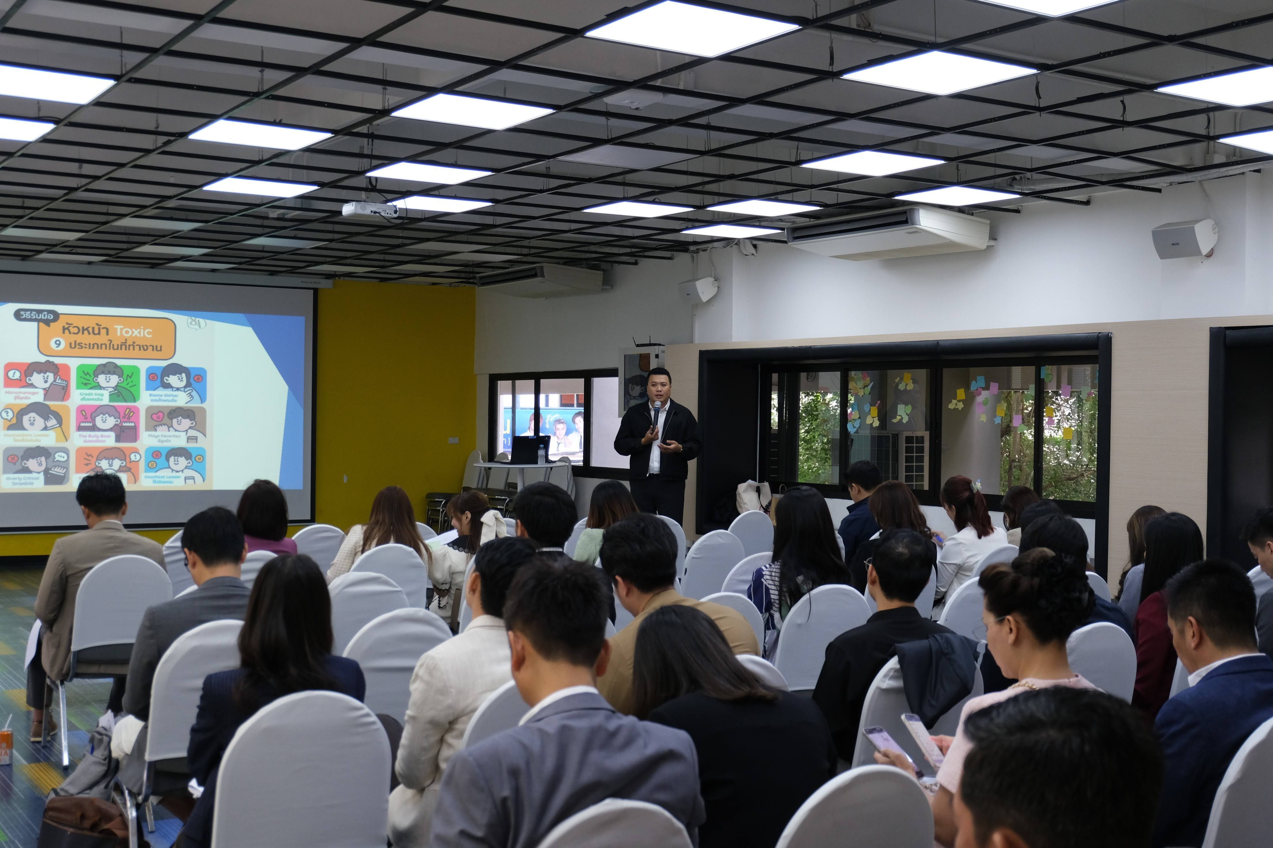 A speaker presents to an audience seated in rows, with a screen displaying content.