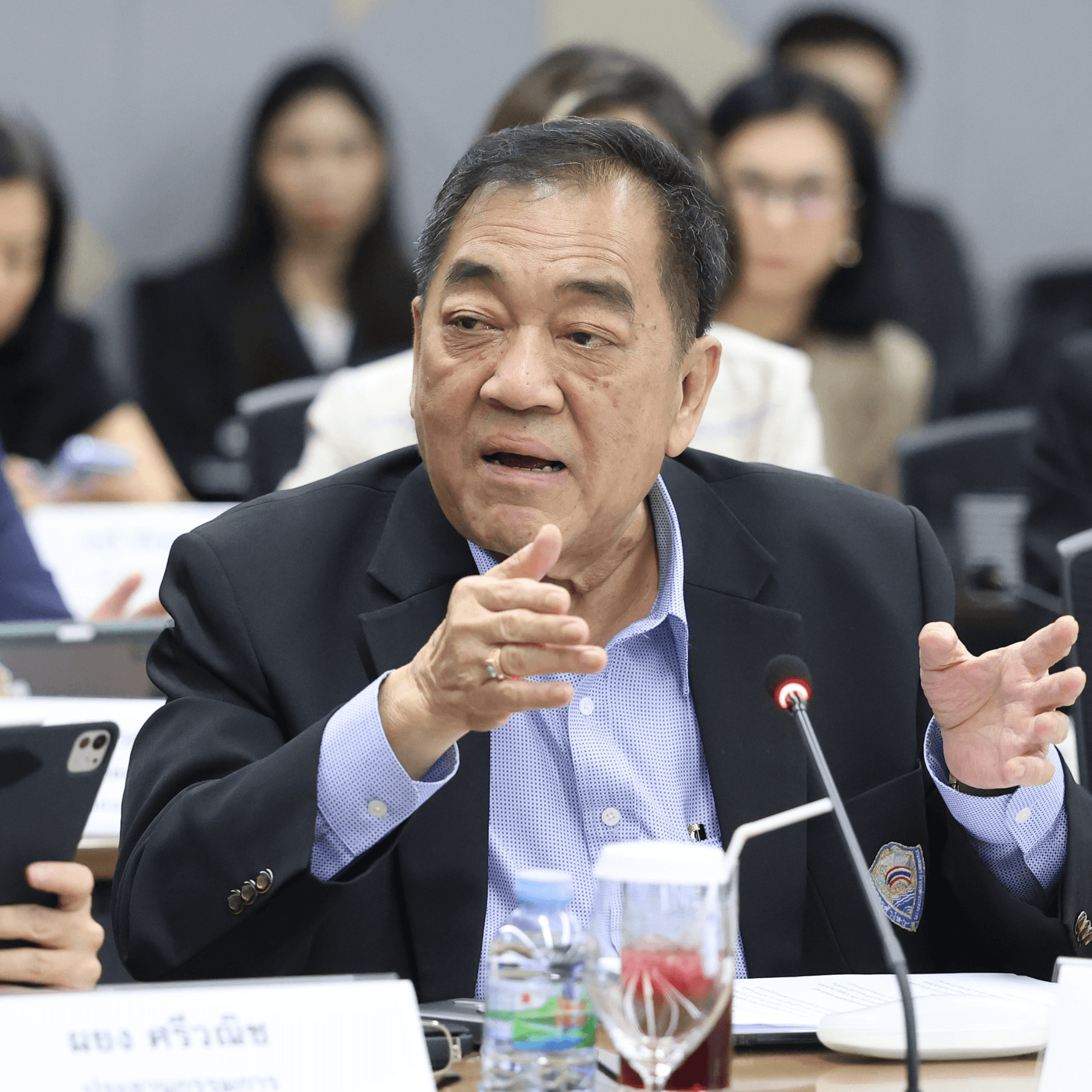 Older man in dark suit speaks and gestures at a professional meeting table.