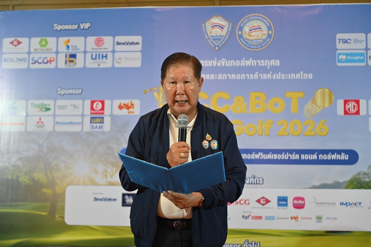 Man in a dark jacket speaks into a microphone at an event with sponsor logos.