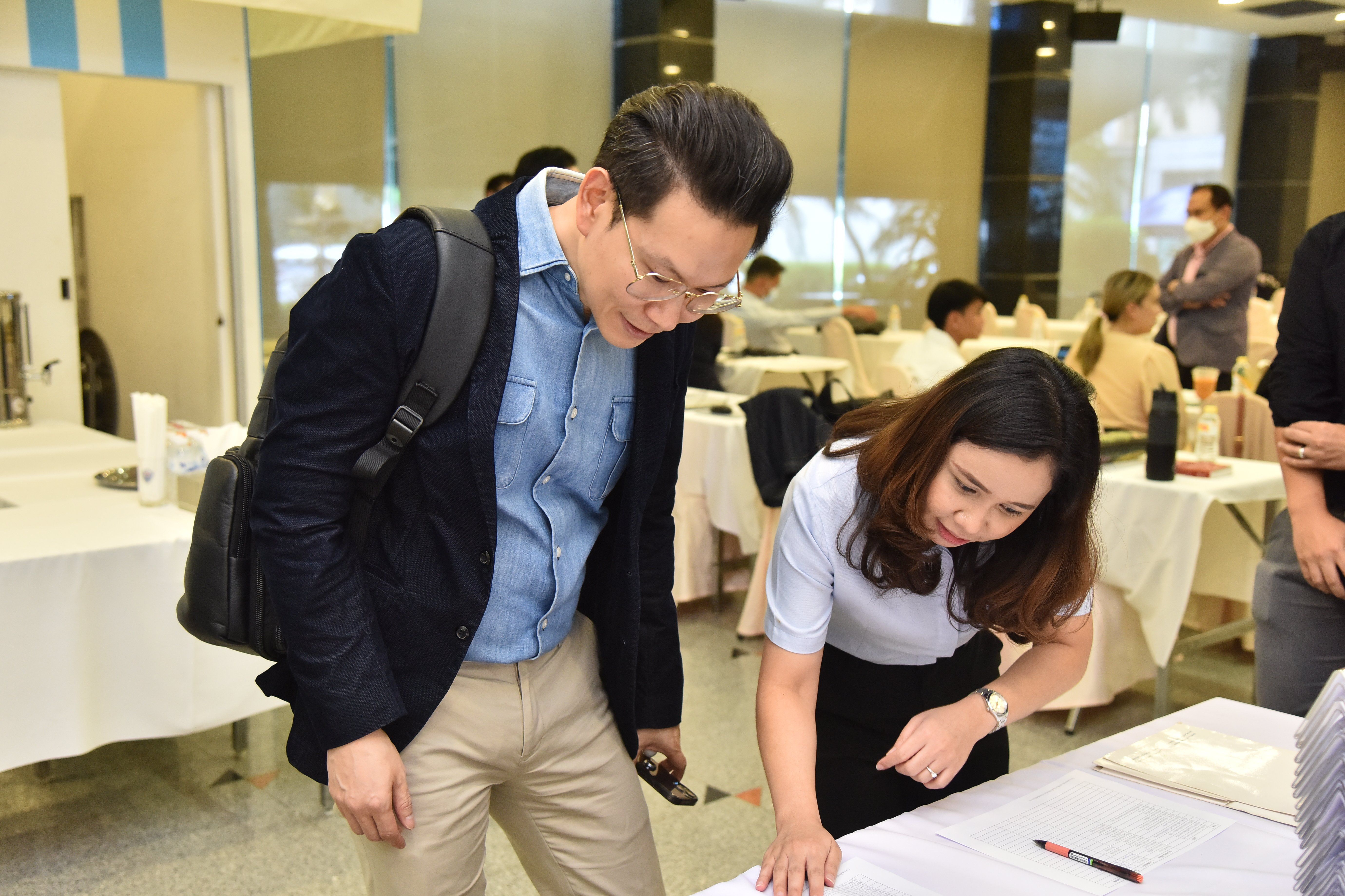 Man watches woman sign documents at an indoor event, other attendees are in background.