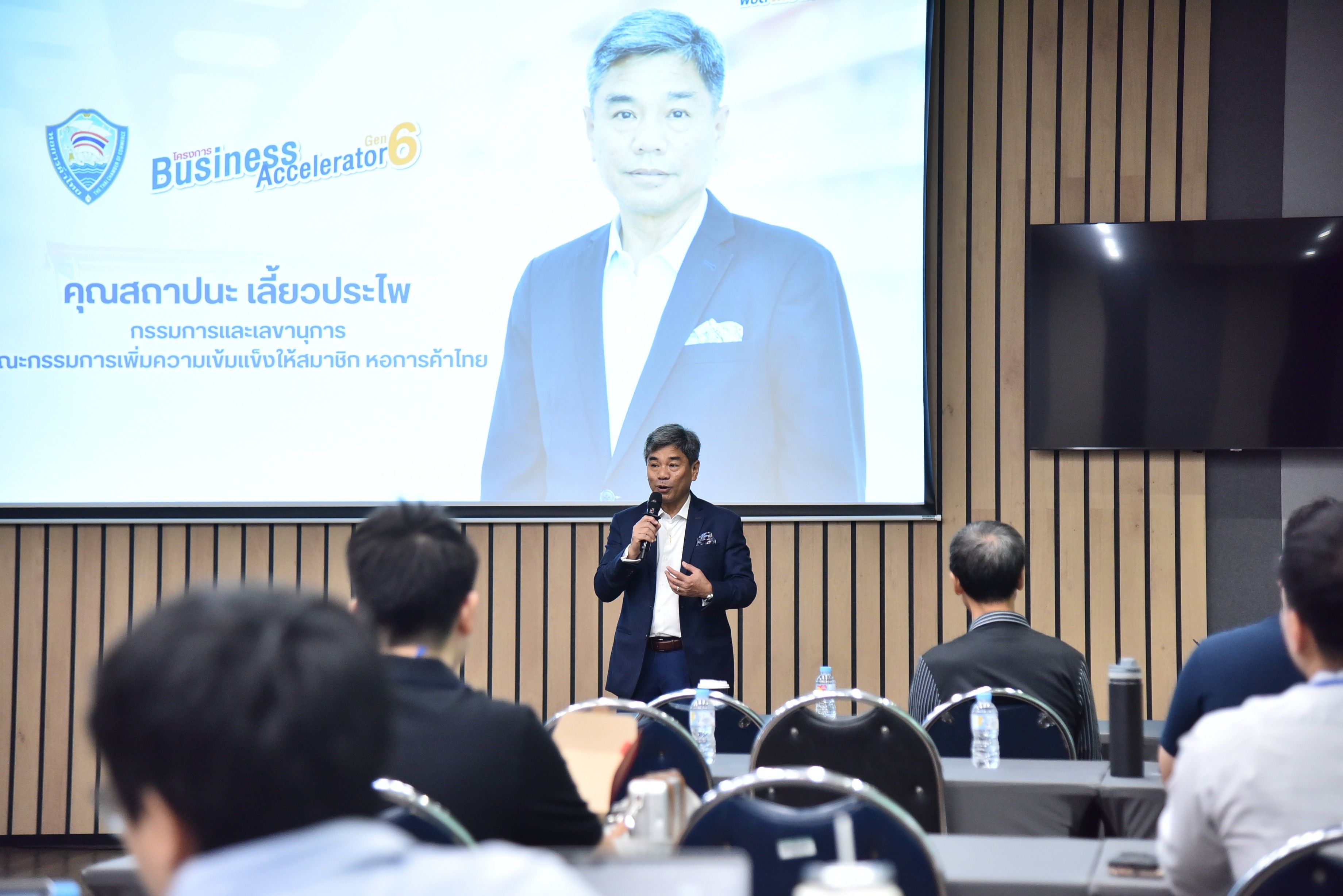 Man in suit speaks at conference with presentation screen and seated audience.