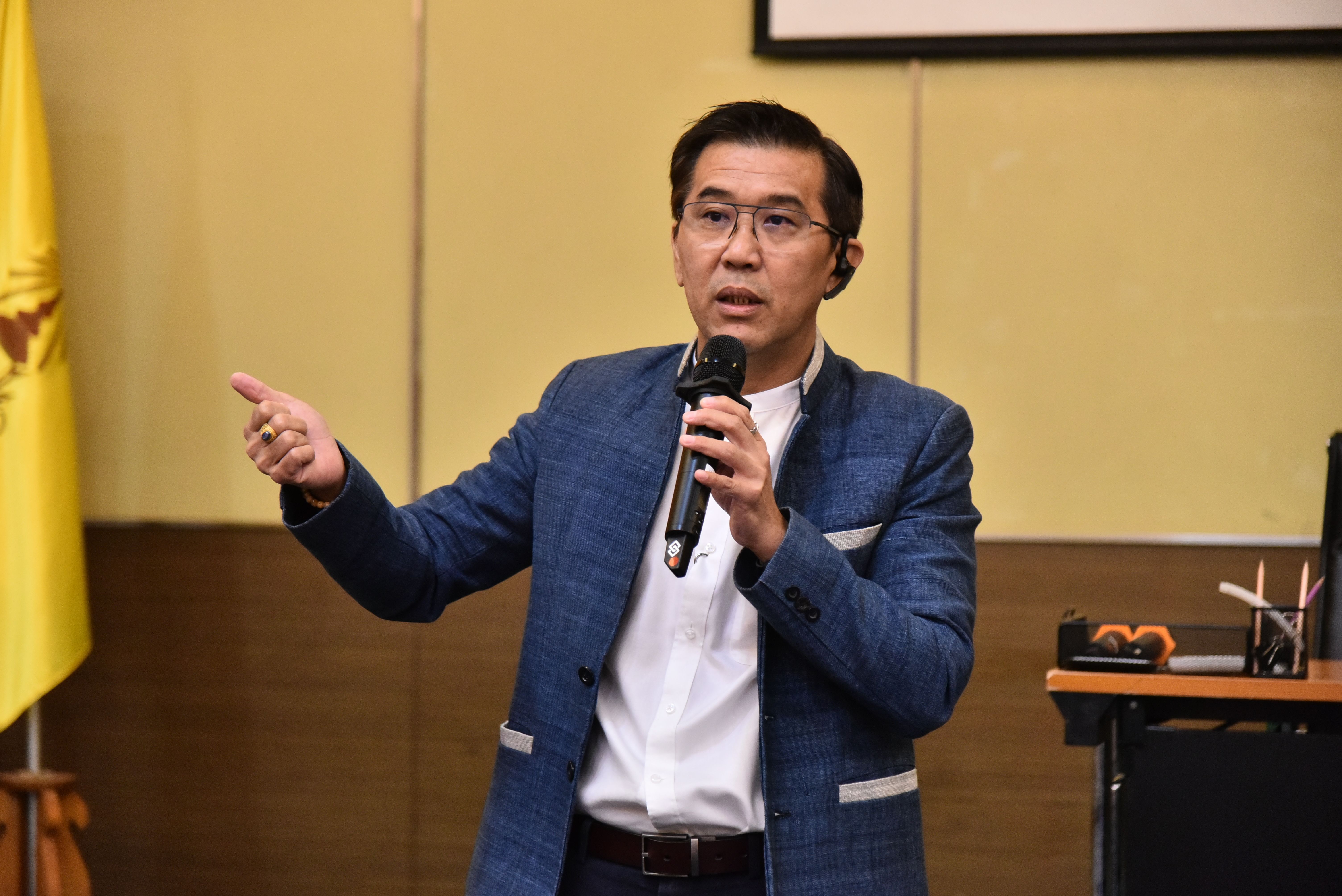 Man in a blue blazer and glasses speaking into a microphone, gesturing.