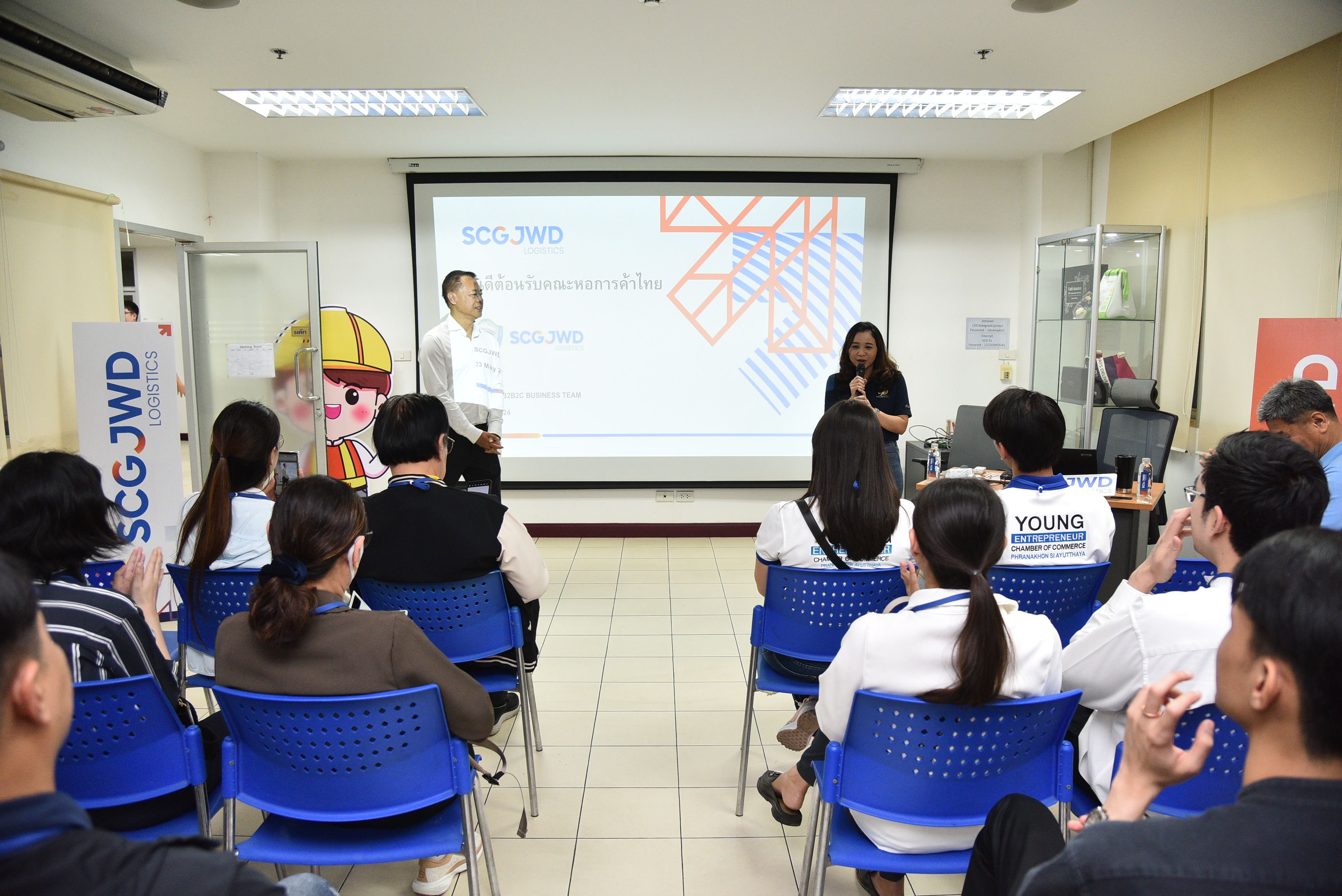 An audience attends a presentation with two speakers and a projector screen displaying 'SCG JWD' logo.