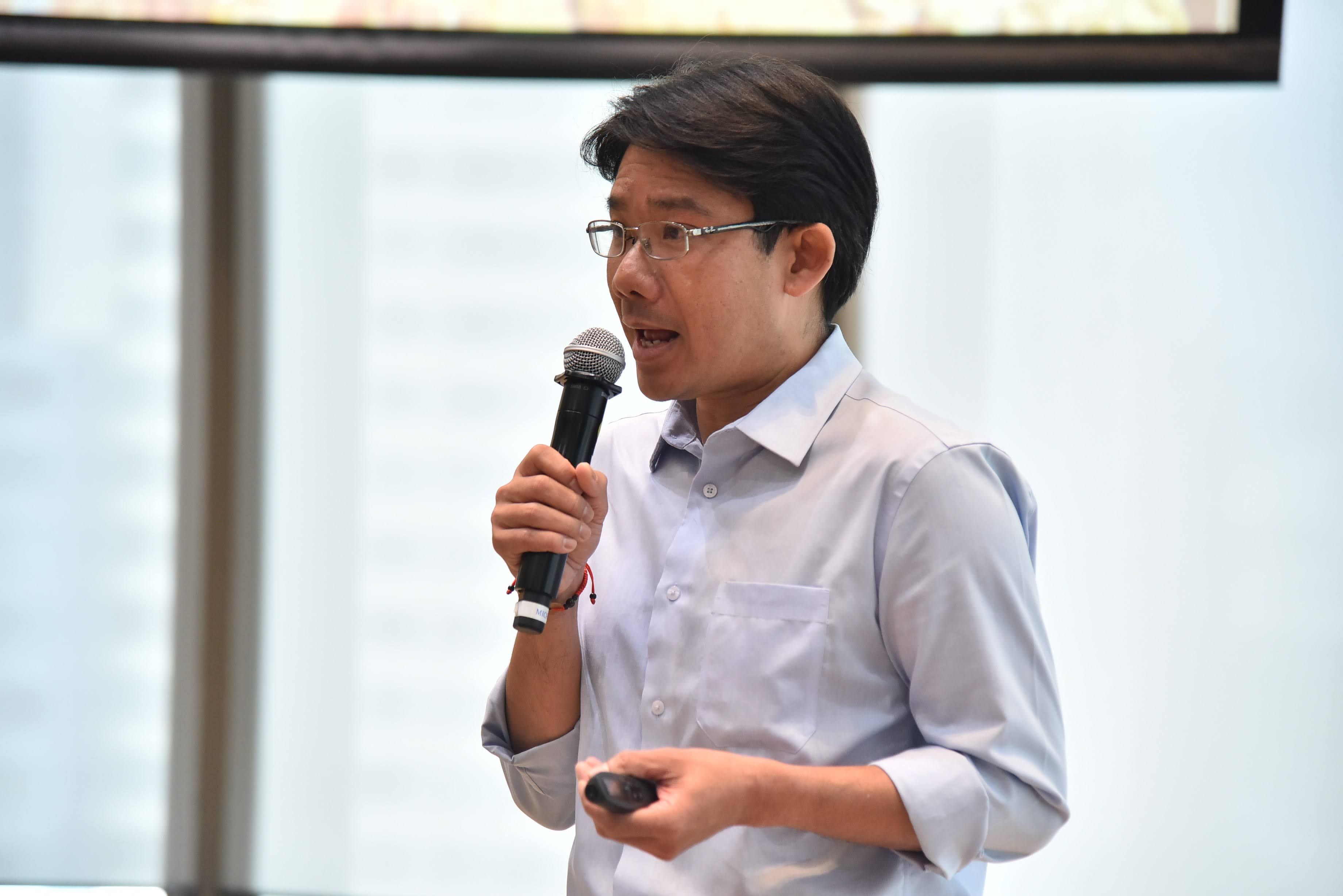 Man in glasses speaking into a microphone, holding a presentation clicker.