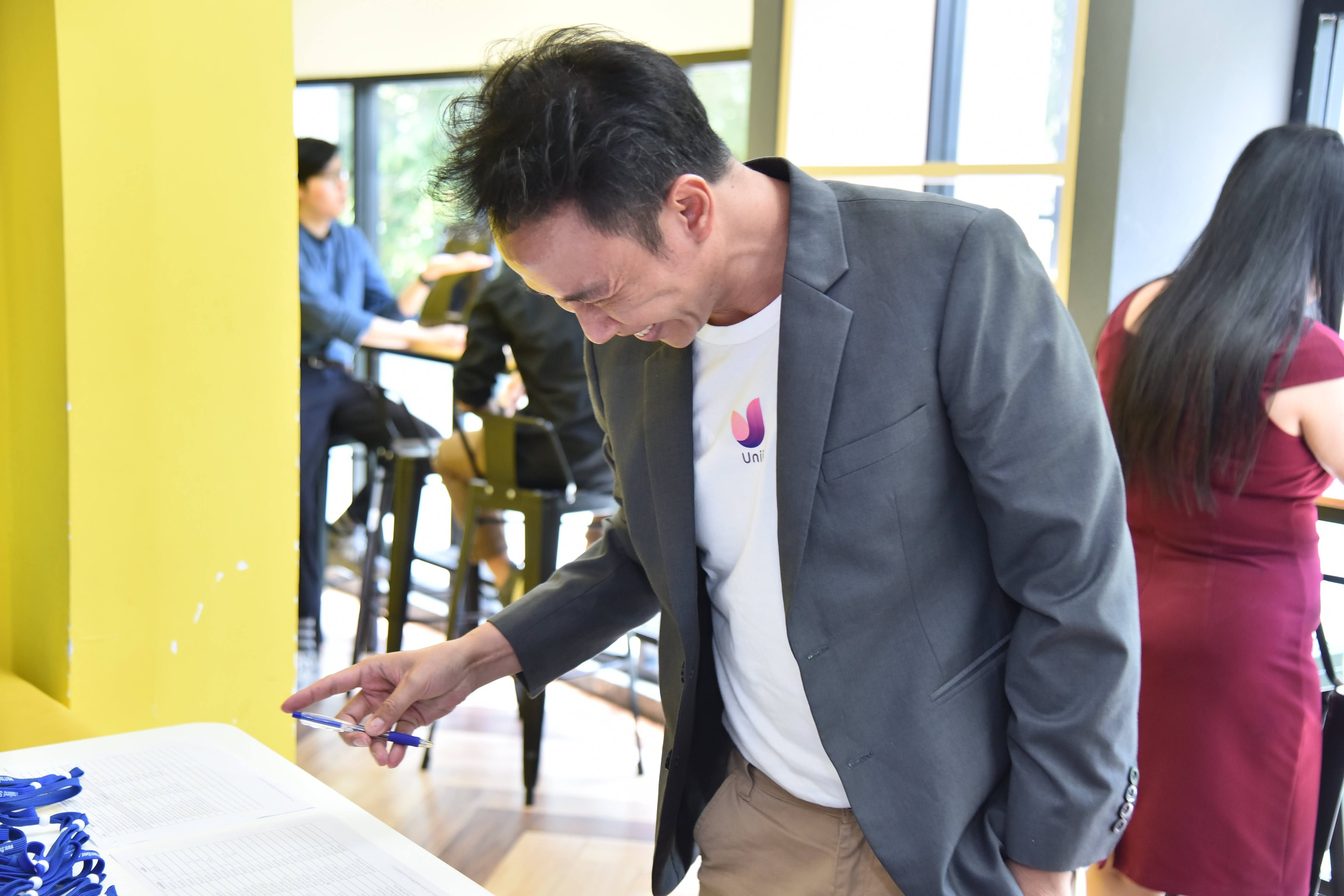 Man in gray jacket points at blue items on a white table in a bright room.