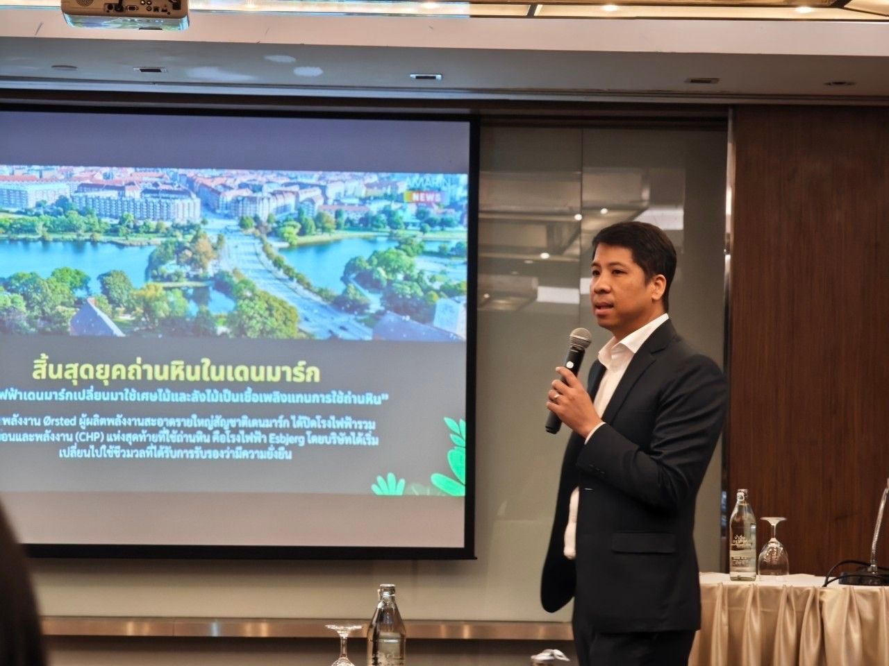Man in suit speaking into microphone during a presentation with a city landscape on screen.
