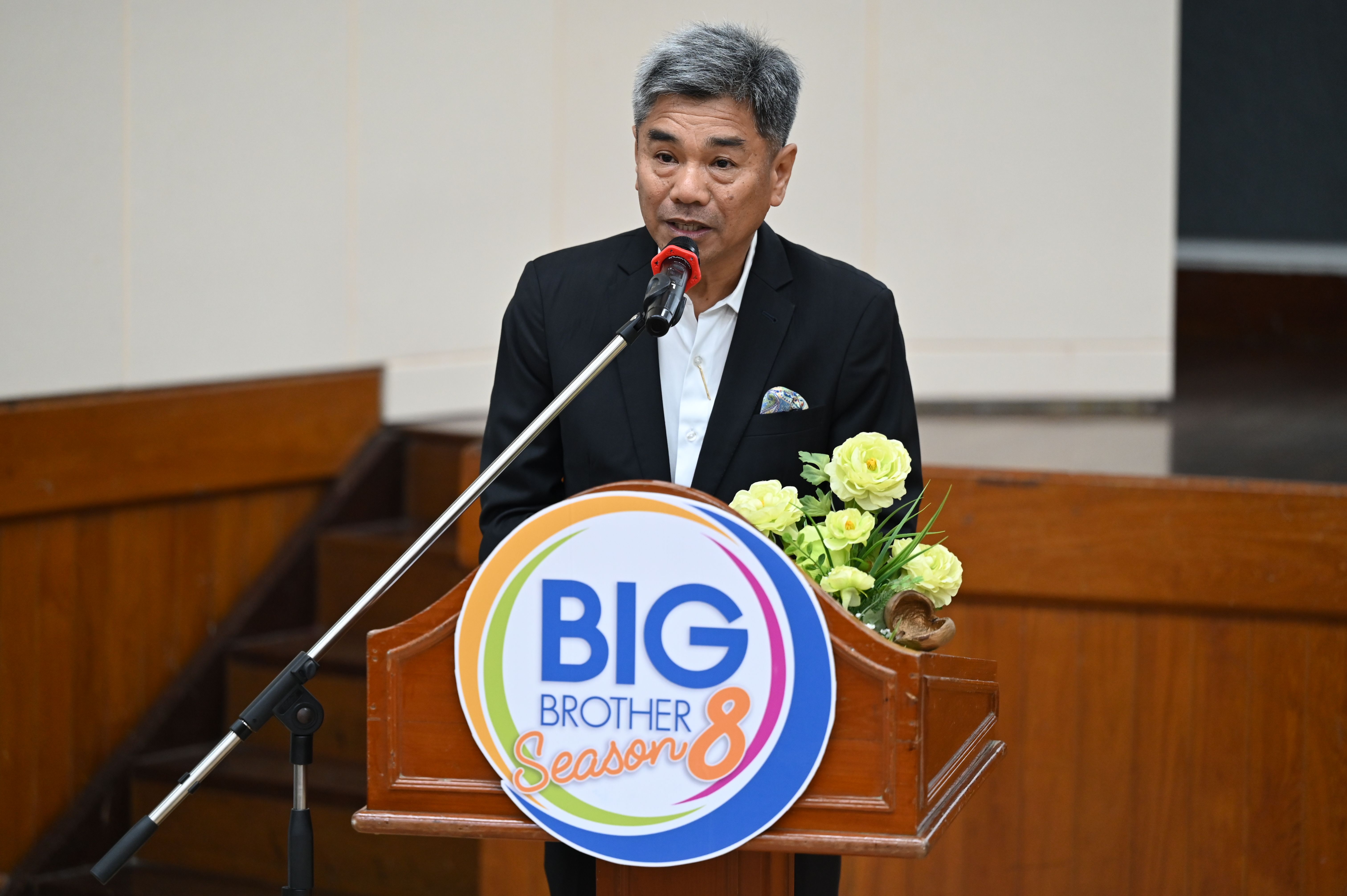 A man speaks at a podium featuring a 'BIG BROTHER Season 8' sign and flowers.
