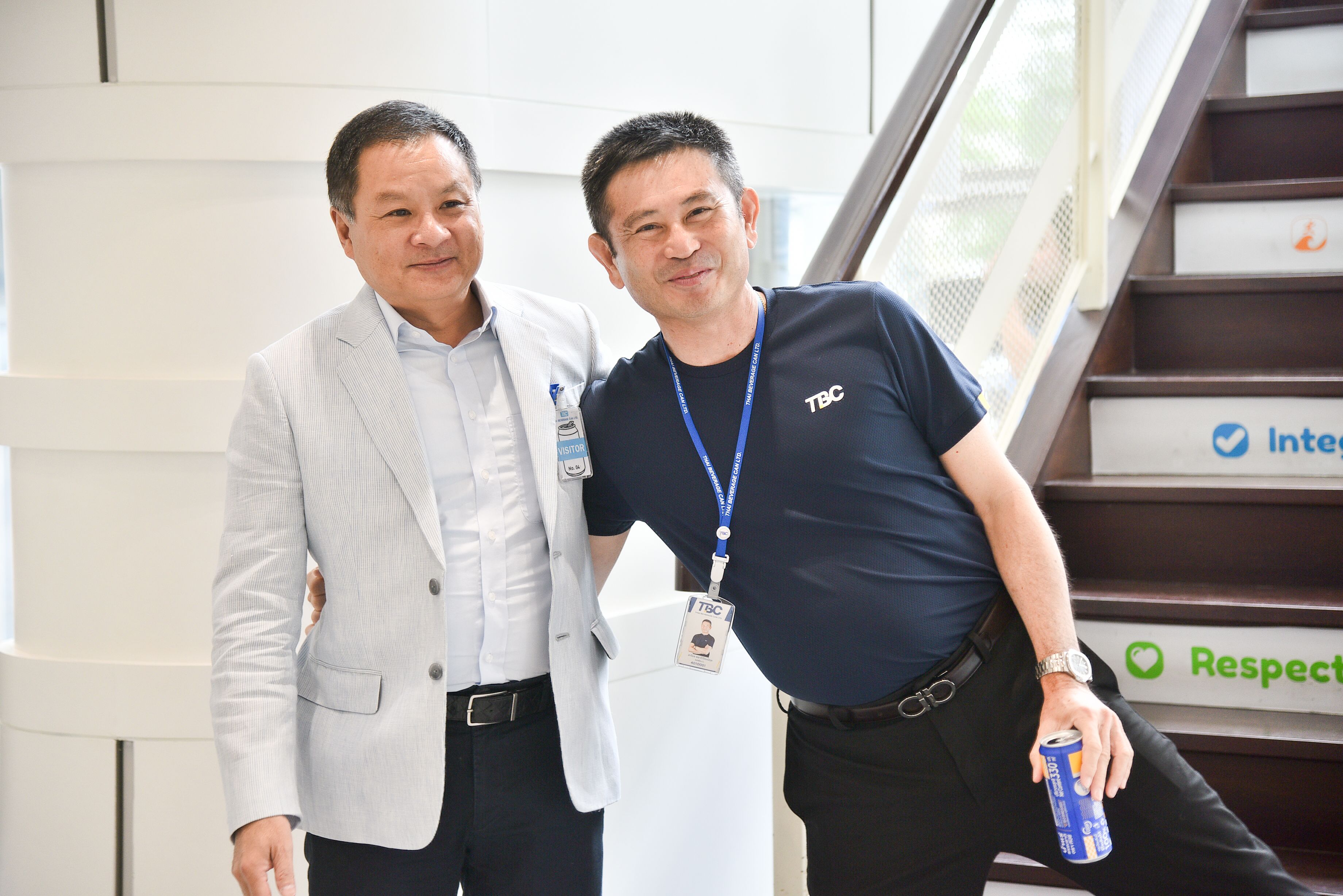 Two men, one in a light suit and one in a dark t-shirt, smiling indoors.