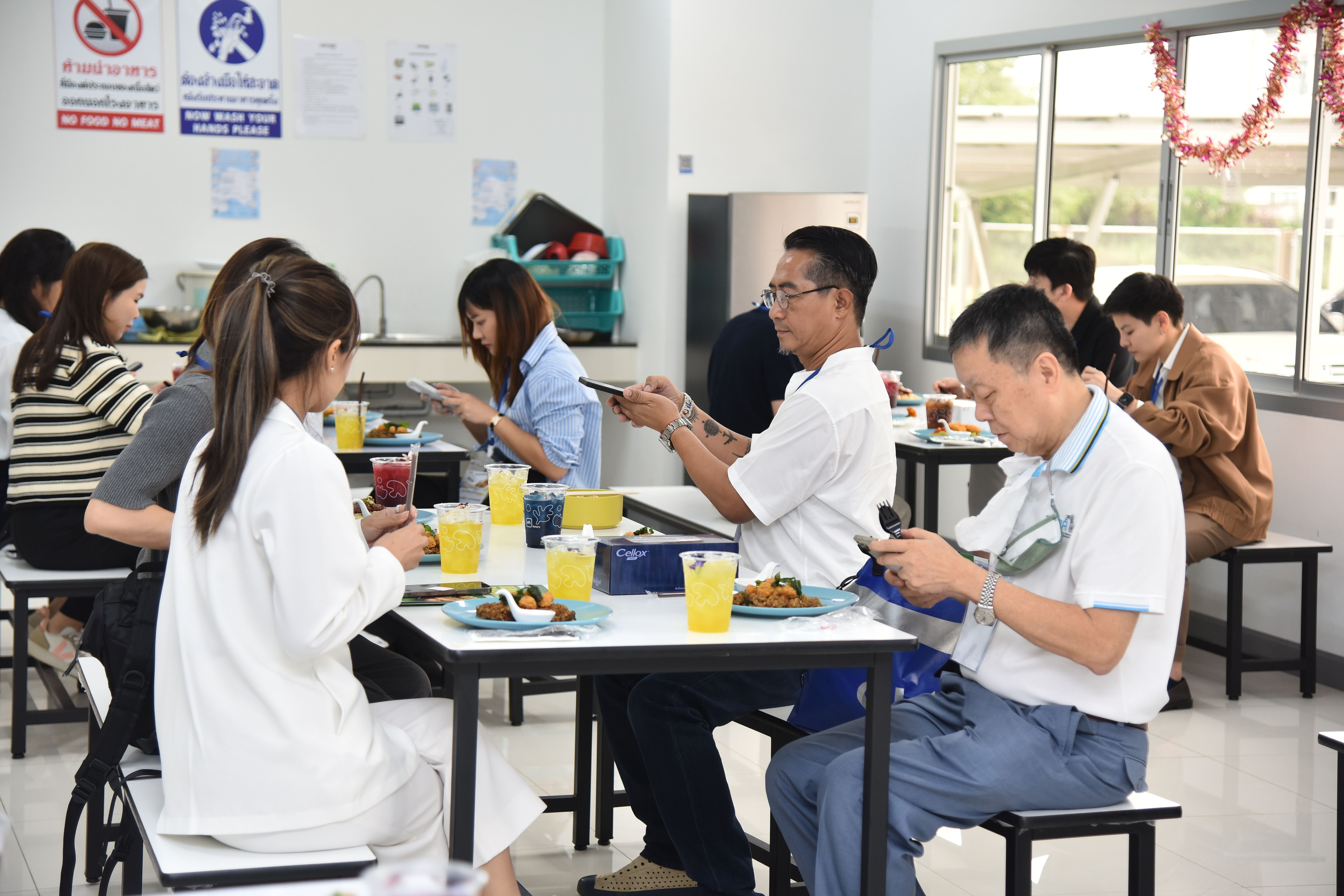 People eating lunch at tables in a cafeteria, many engrossed in their smartphones.