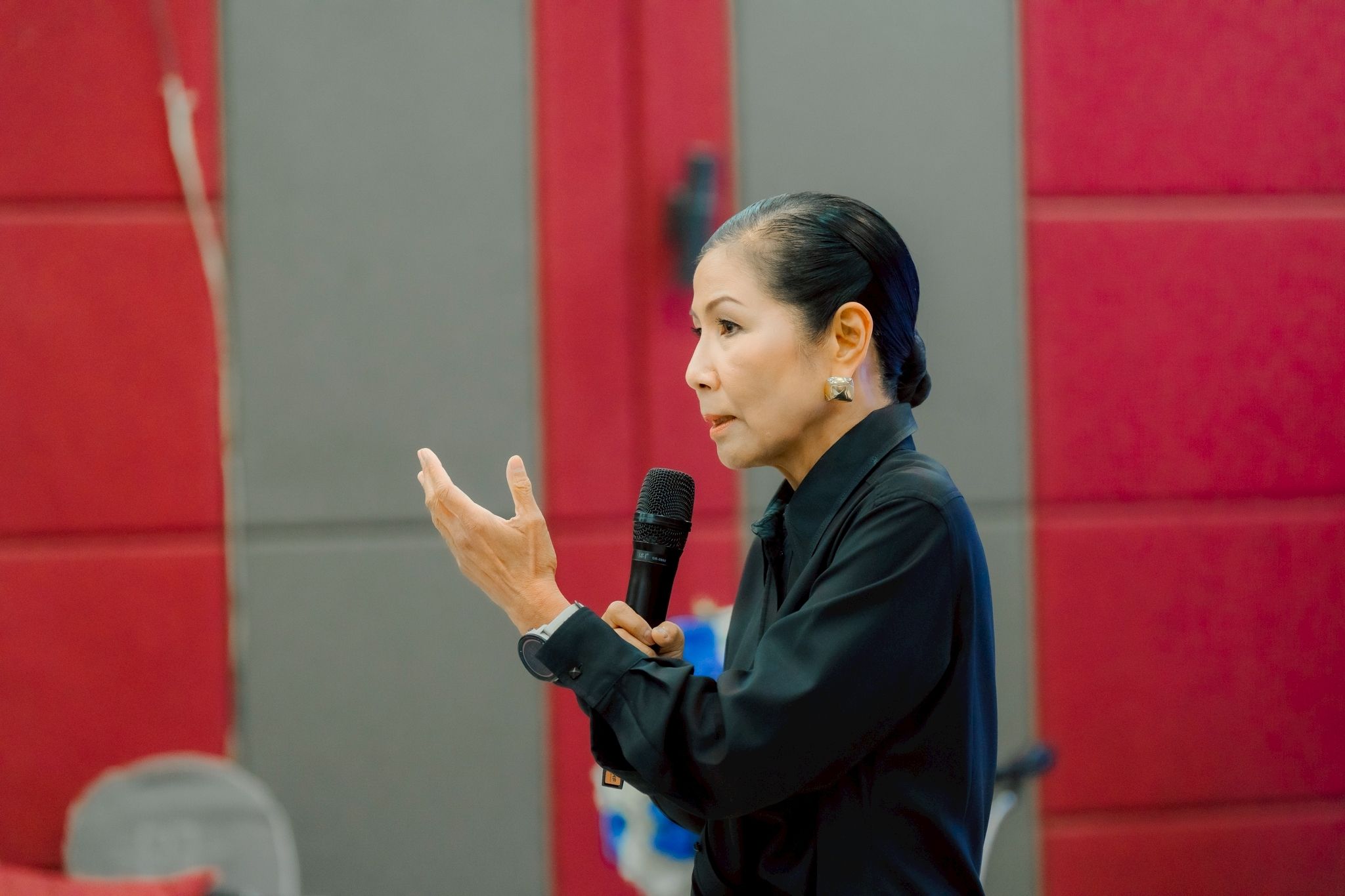 A woman in a black shirt speaks into a microphone, gesturing with her hand.