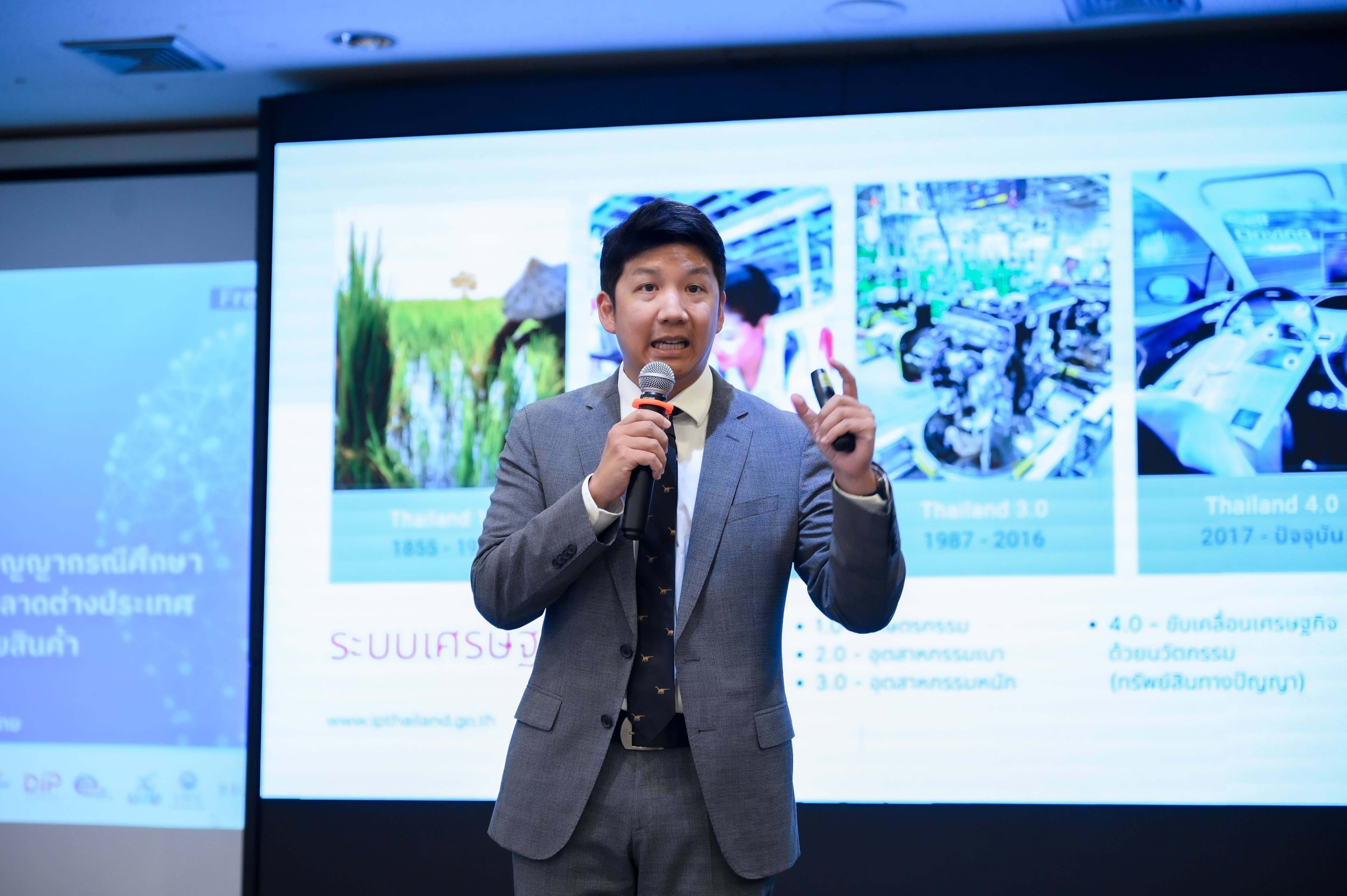 Man in a grey suit speaking into a microphone during a presentation with visual slides.