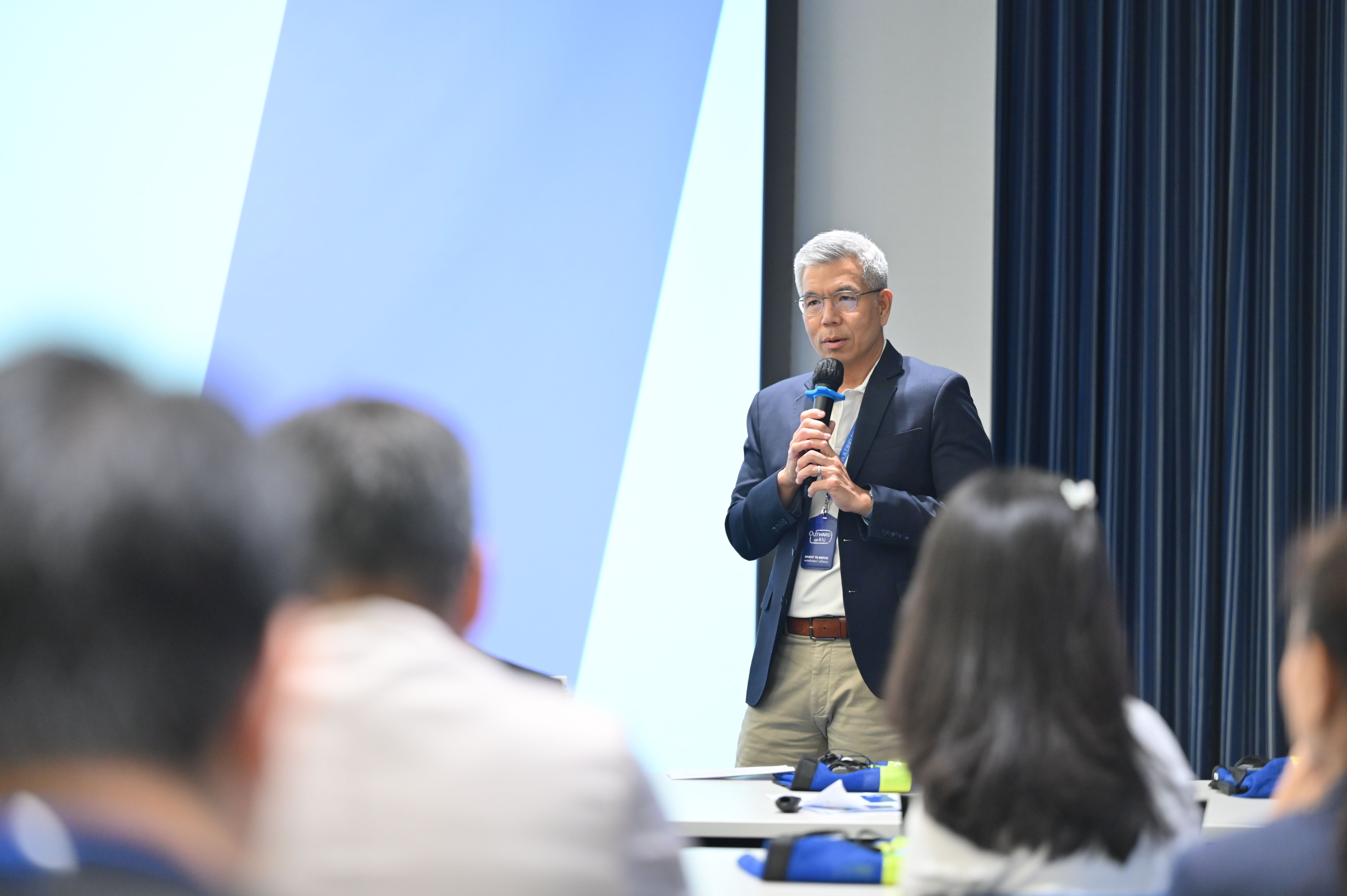 Man speaking into a microphone at a conference in front of a blue screen.