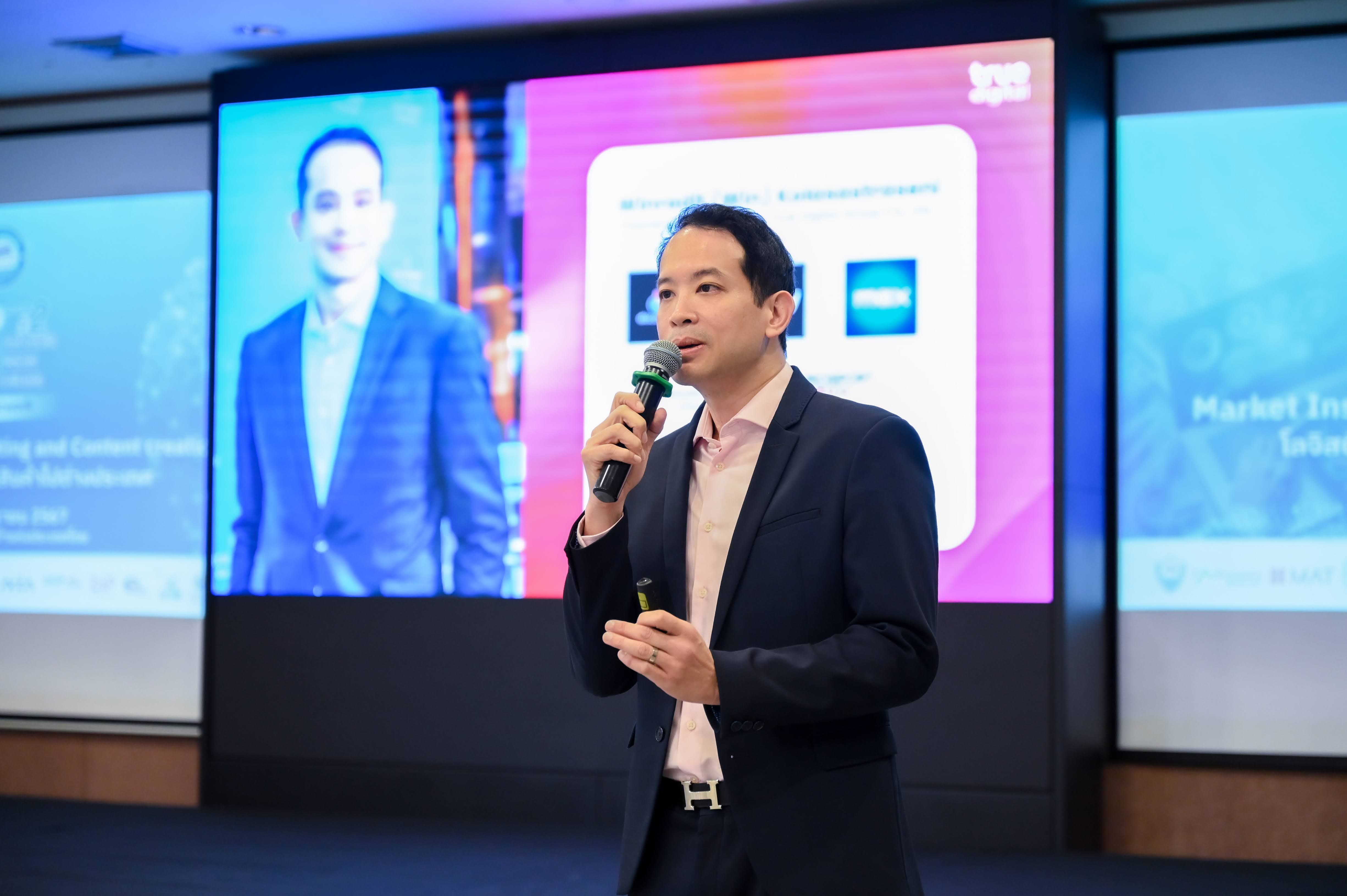 A professional speaker presents on stage, holding a microphone, with digital screens behind him.