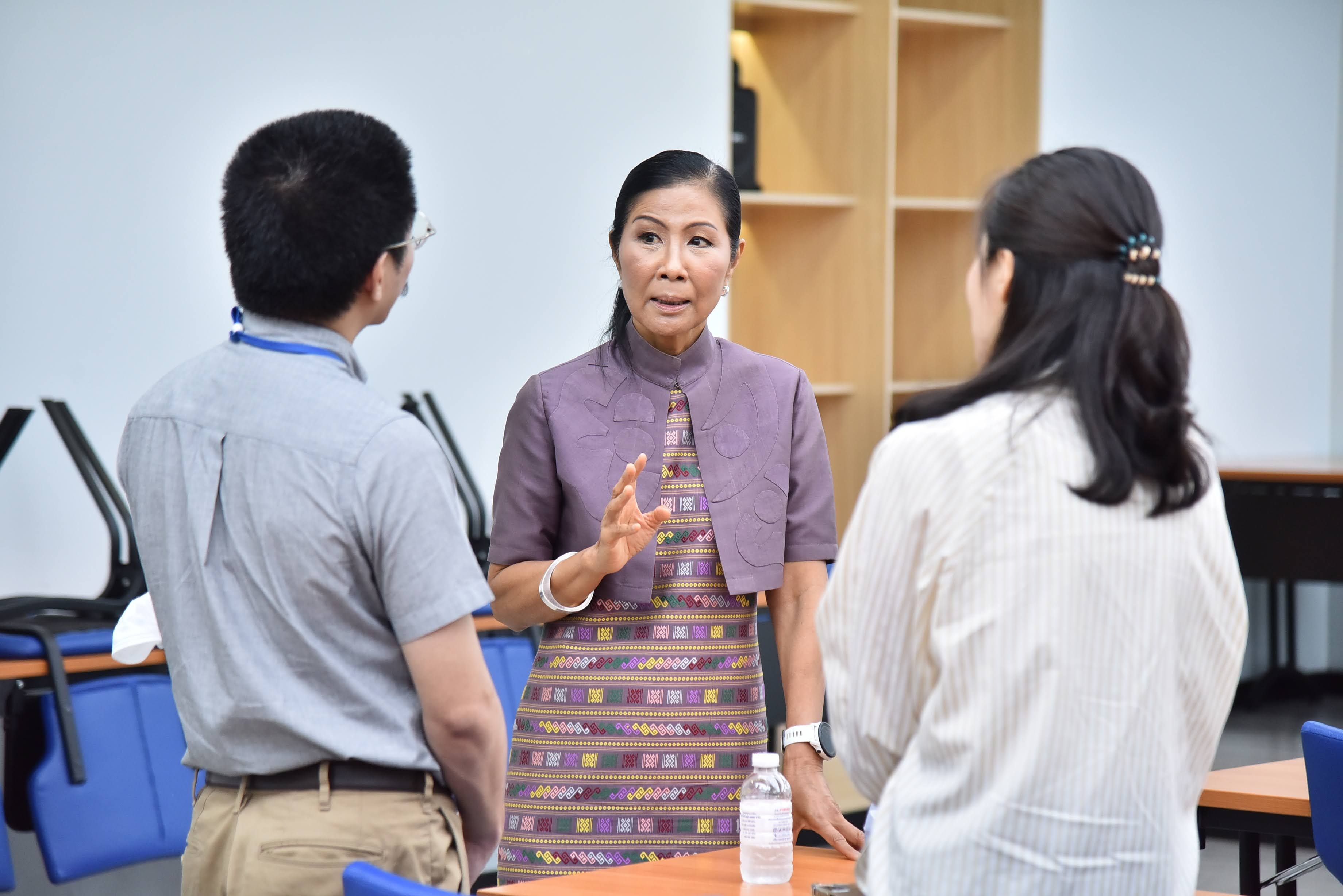 A woman in a purple patterned outfit gestures while speaking to two individuals in a meeting.